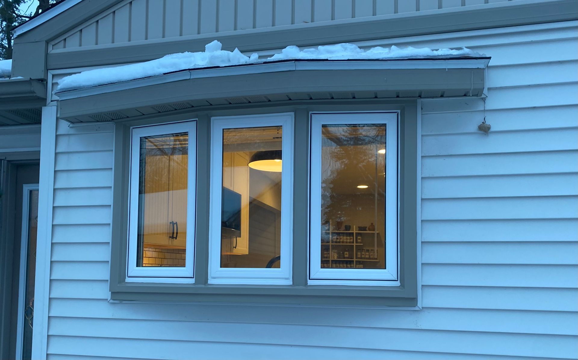 Blue house exterior with three lit windows and snow on the roof at dusk