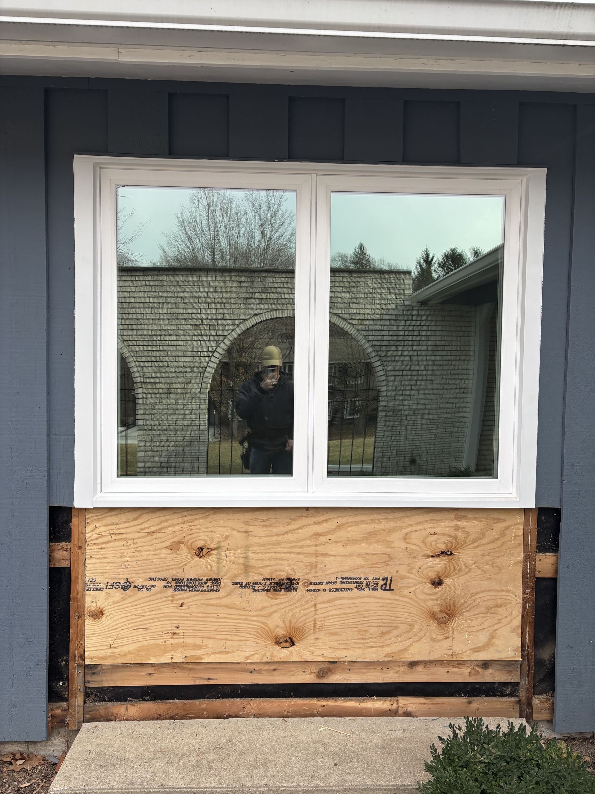 Boarded-up window on a dark blue exterior wall, with a large white-framed glass window above plywood.