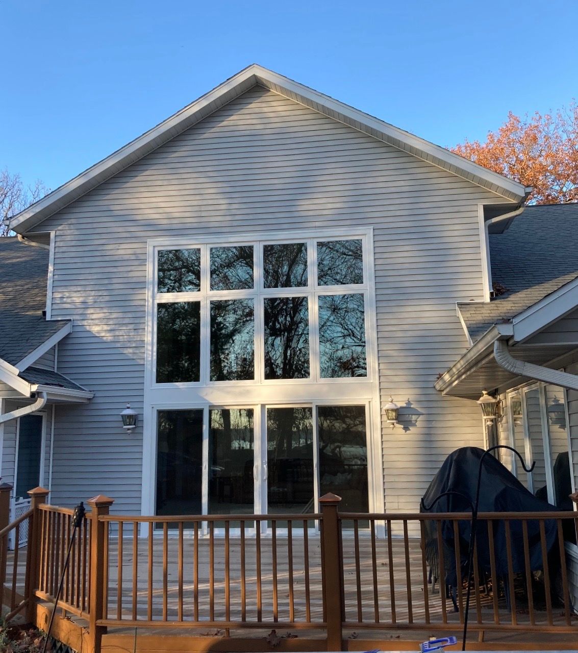 Back of a suburban house with tall windows, wooden deck, and a covered grill in the yard