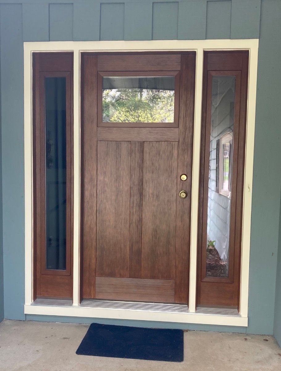 Front porch with a brown wooden door, narrow glass sidelights, and a blue doormat against a gray wall