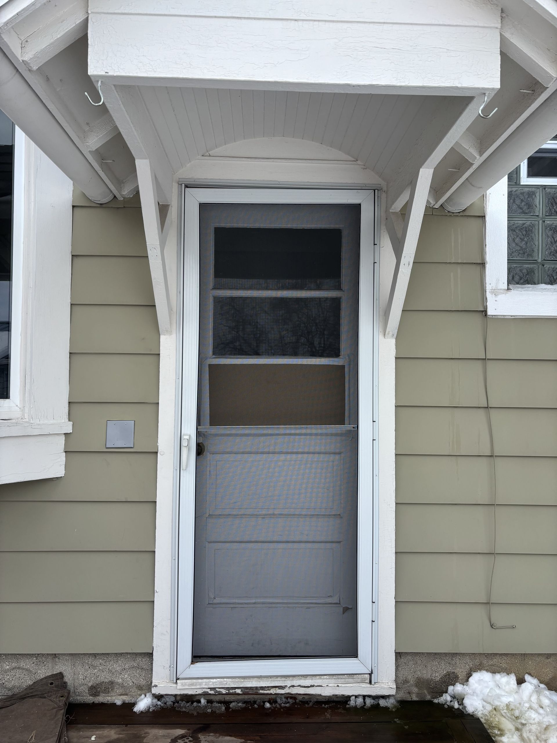 Snow-covered front entrance with gray door, white trim, and small awning on a beige house