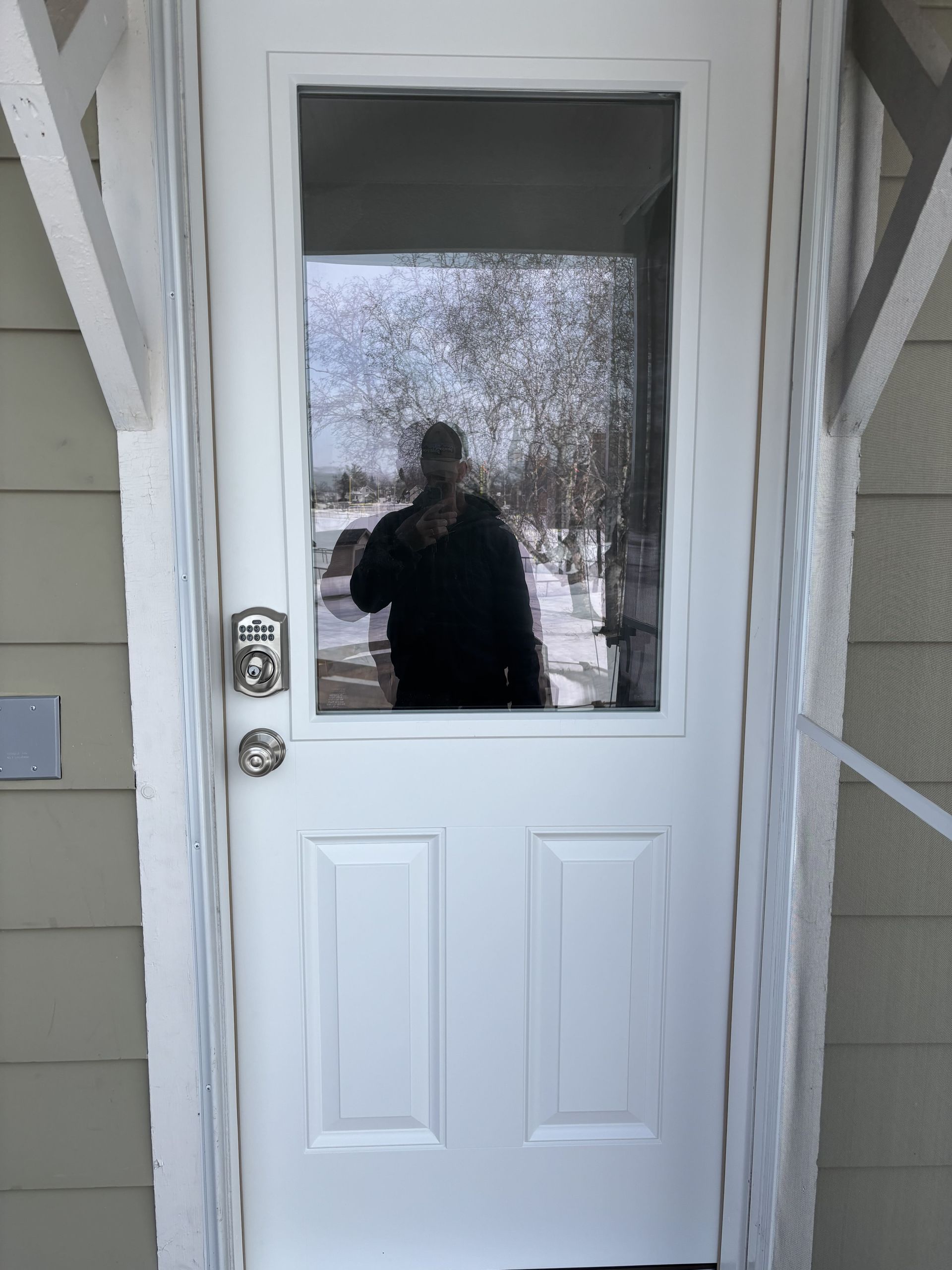 White front door with window reflecting a person standing outside and snowy trees beyond