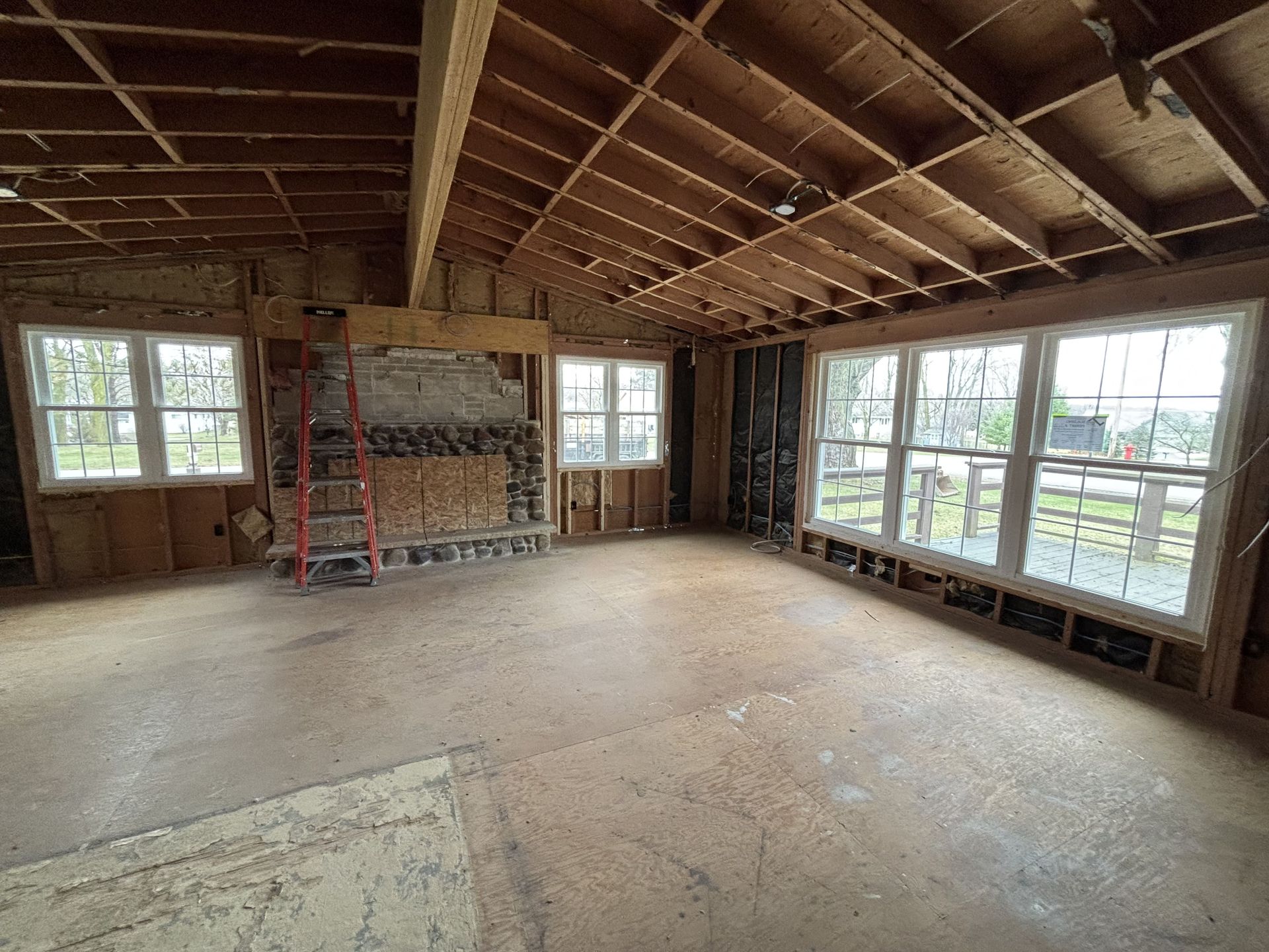 Unfinished room with exposed ceiling beams, open windows, a ladder, and a stone fireplace.