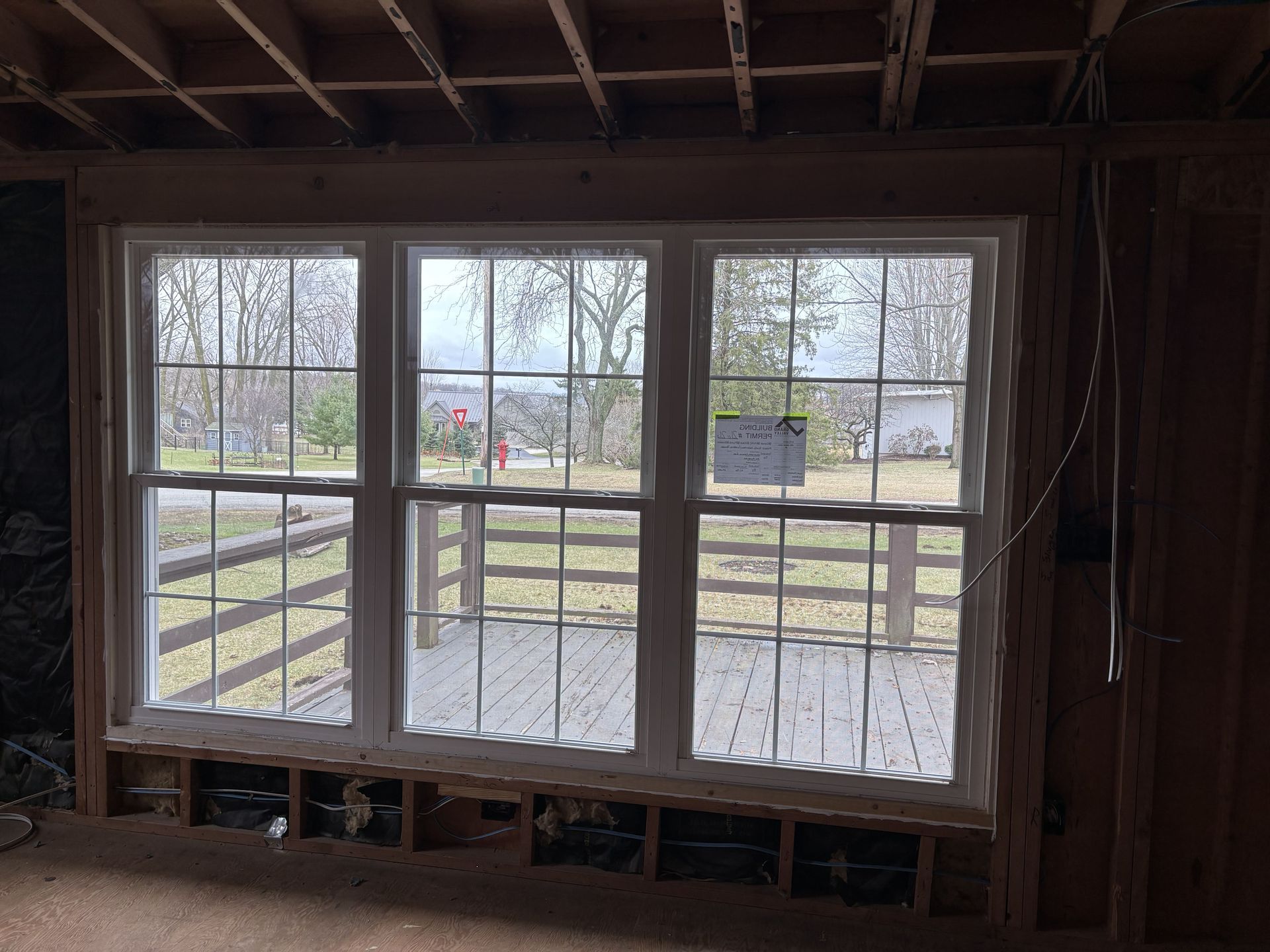 Large window framed in unfinished wall, overlooking a backyard and road on a cloudy day