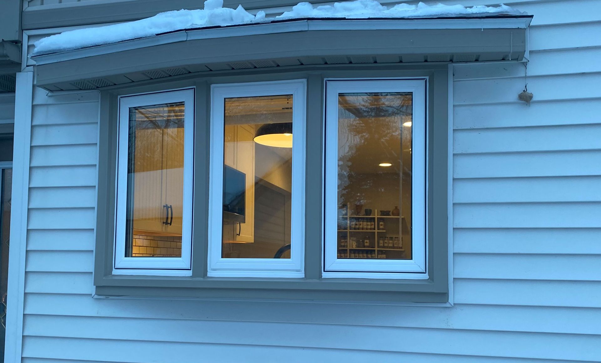 White bay window with blinds on a gray-sided house exterior under the eaves