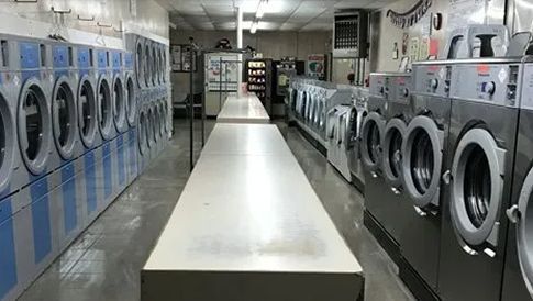 Interior of a laundromat. Rows of washing machines face a long, white folding table in a brightly lit, industrial setting.