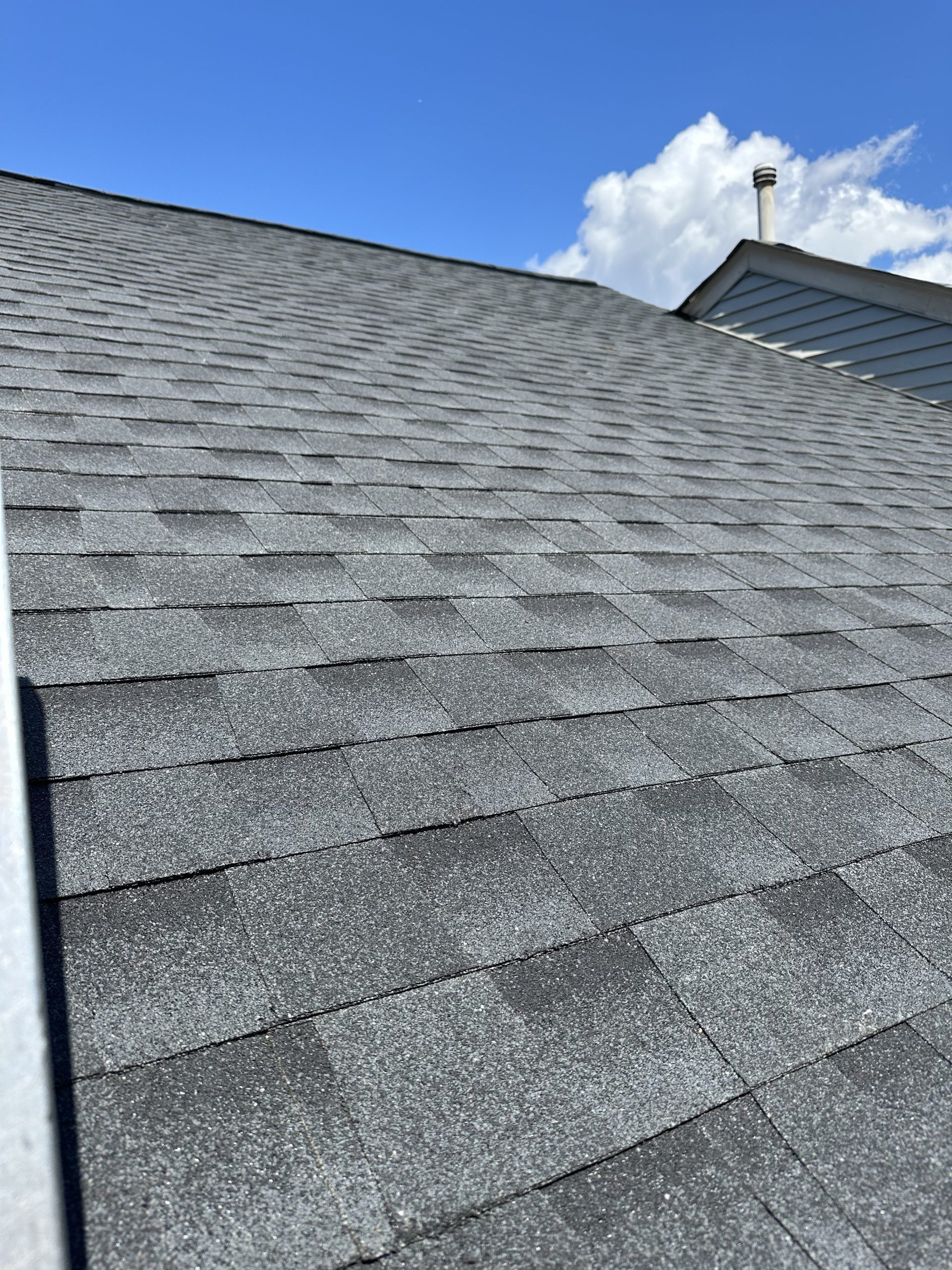 Gray shingled roof under a blue sky with a small vent pipe and nearby roofline