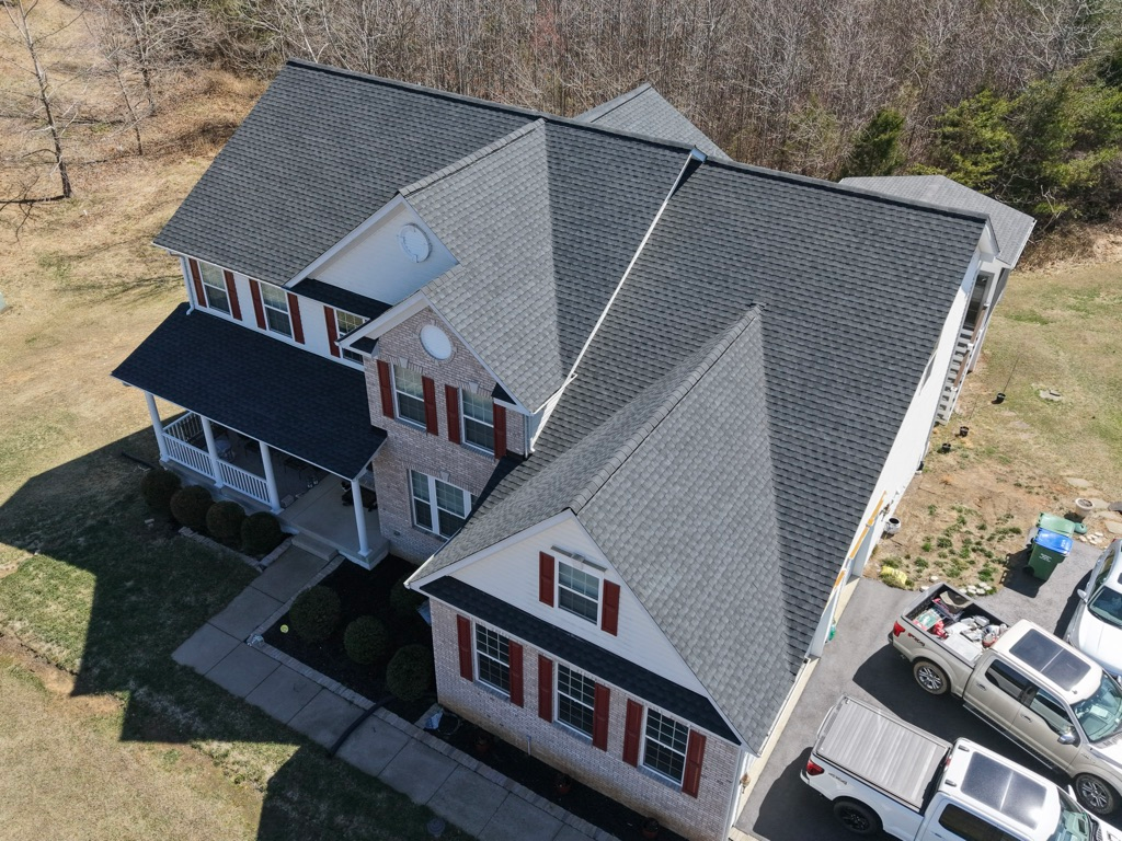 Aerial view of a large gray house with white trim, a black awning, and parked cars beside it.