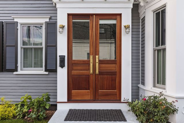 Wooden double front doors with glass panels on a gray house, framed by white trim and black wall lanterns