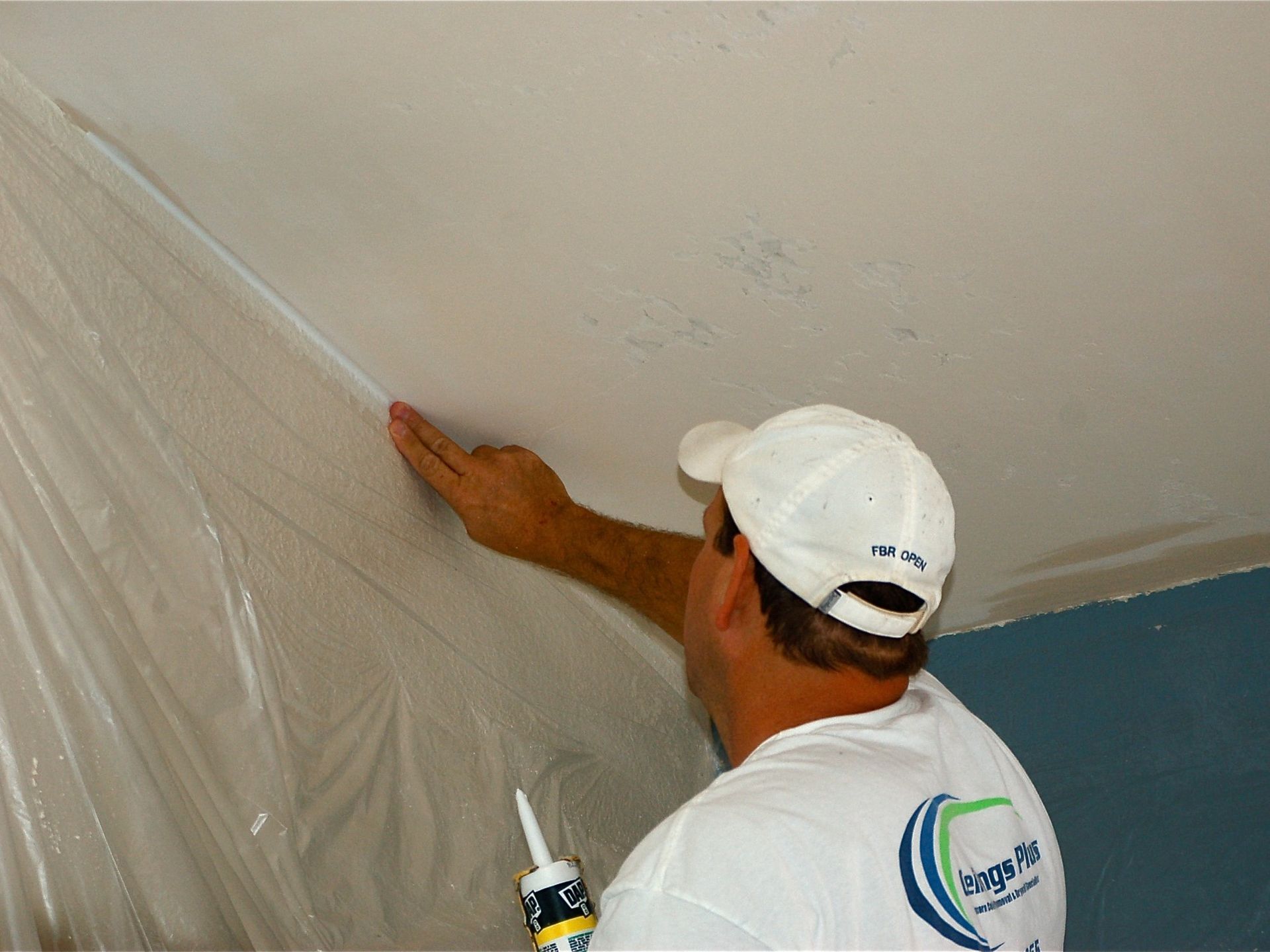 A man in a white hat is applying silicone to a ceiling.