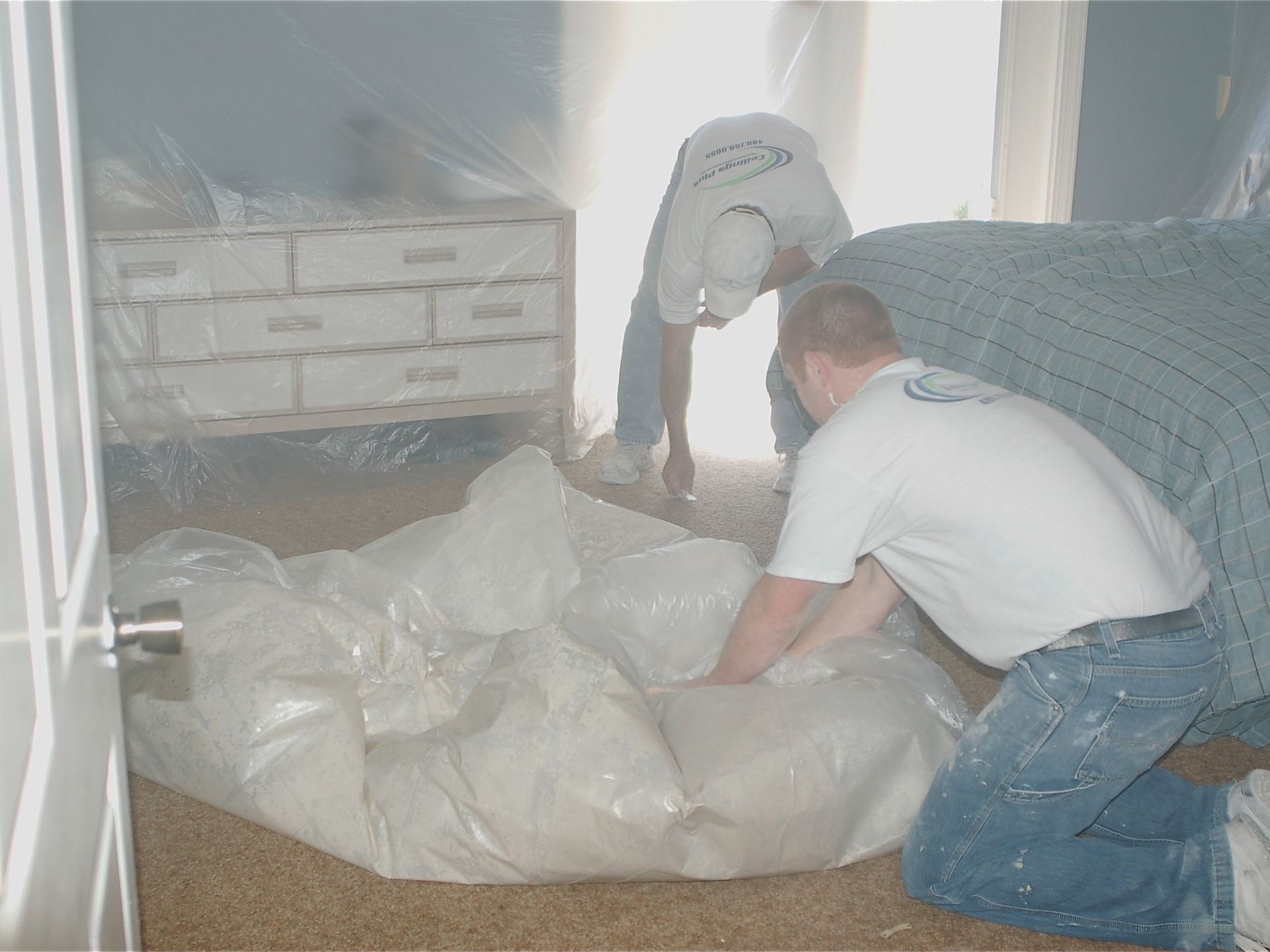 Two men are wrapping a bed in plastic in a bedroom