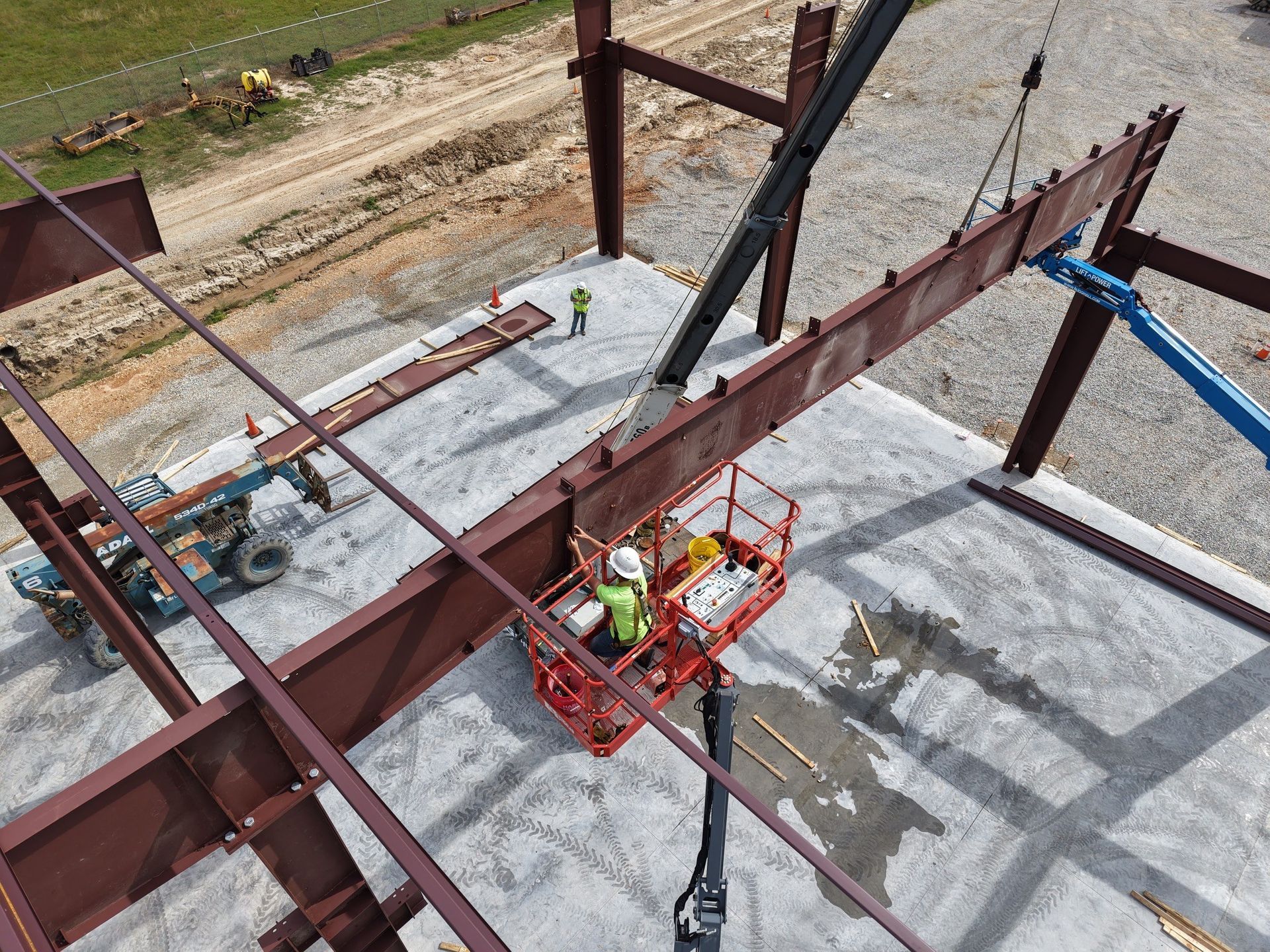 Construction workers in a lift connect a steel beam to a building frame. A crane hoists the beam.