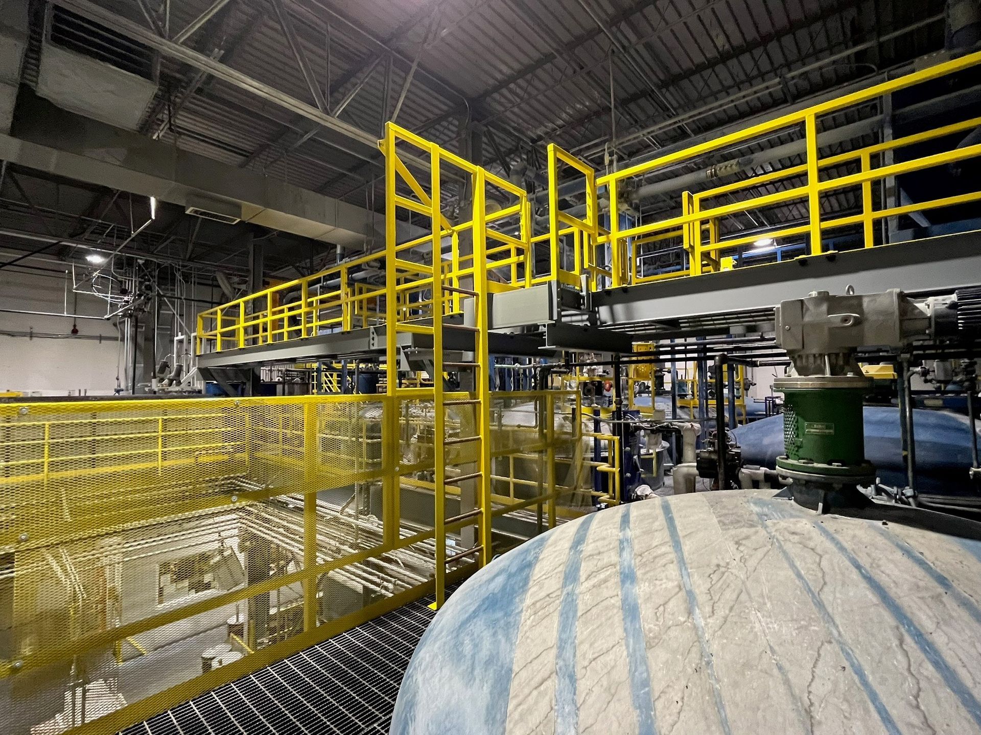 Yellow industrial catwalks and railings in a factory setting, with large gray tanks.