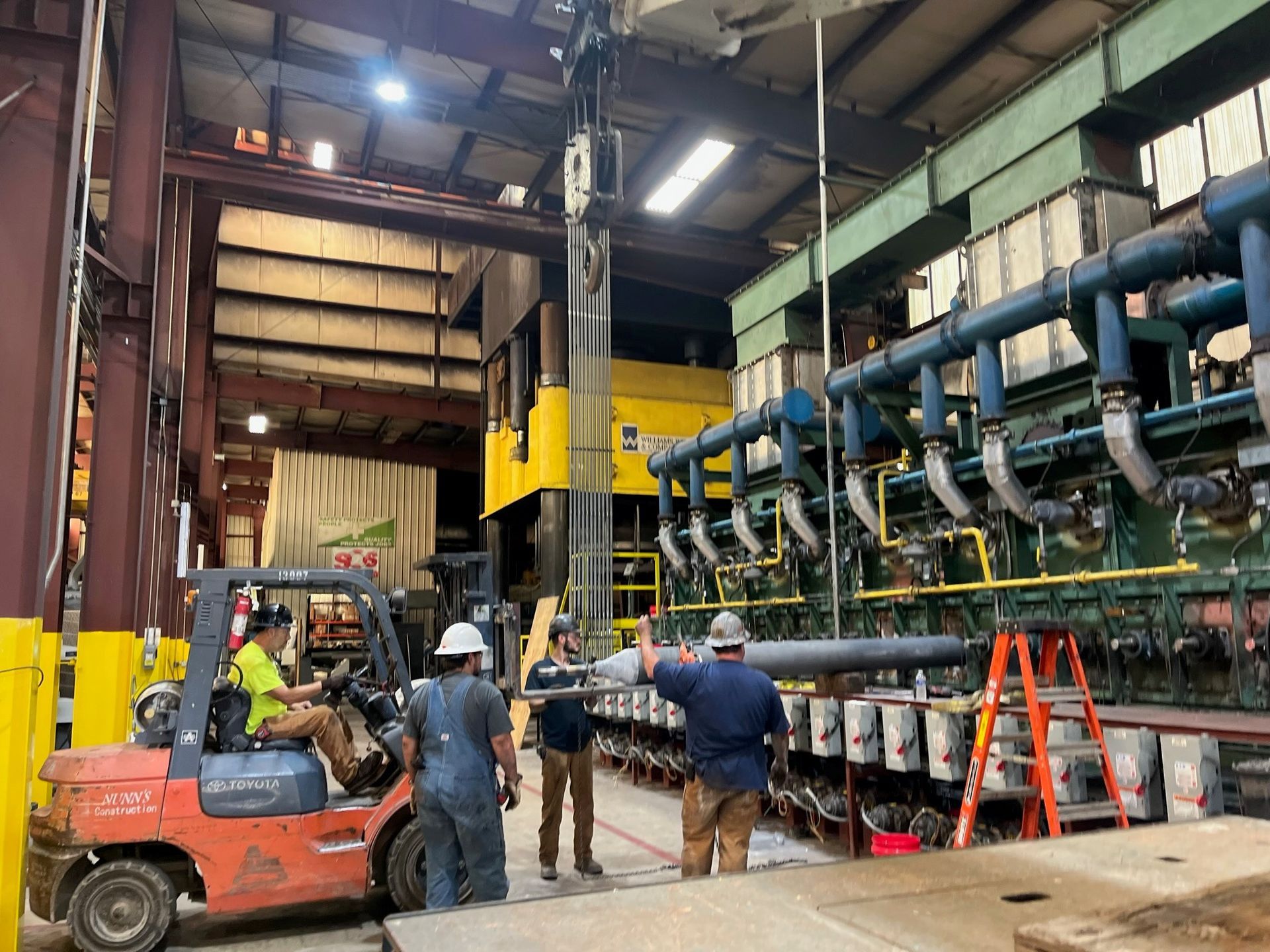 Workers using a forklift and overhead crane to move a long metal cylinder in a factory.