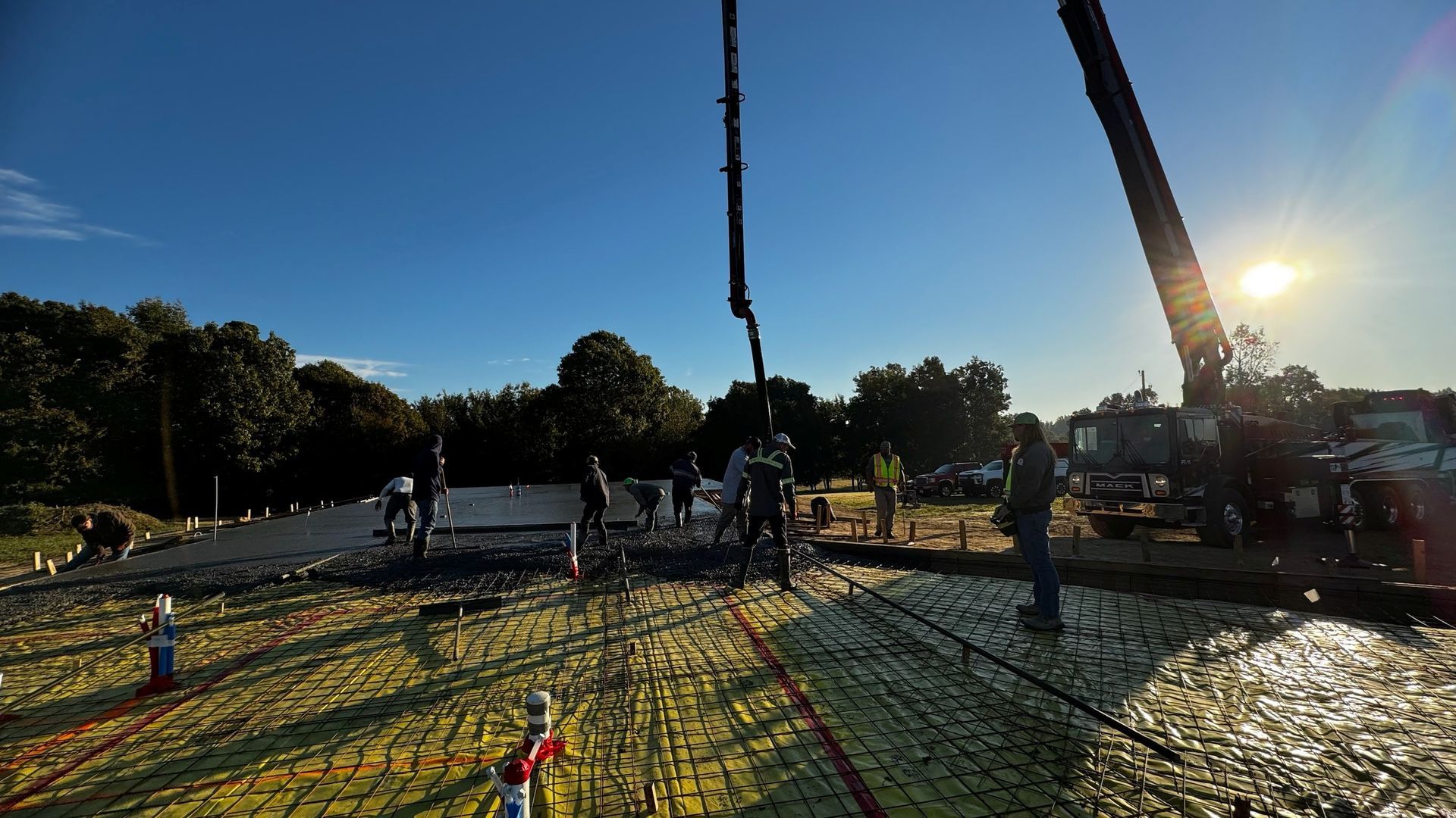Construction crew pouring concrete, using a boom pump, onto a large, yellow insulated foundation under a sunny sky.