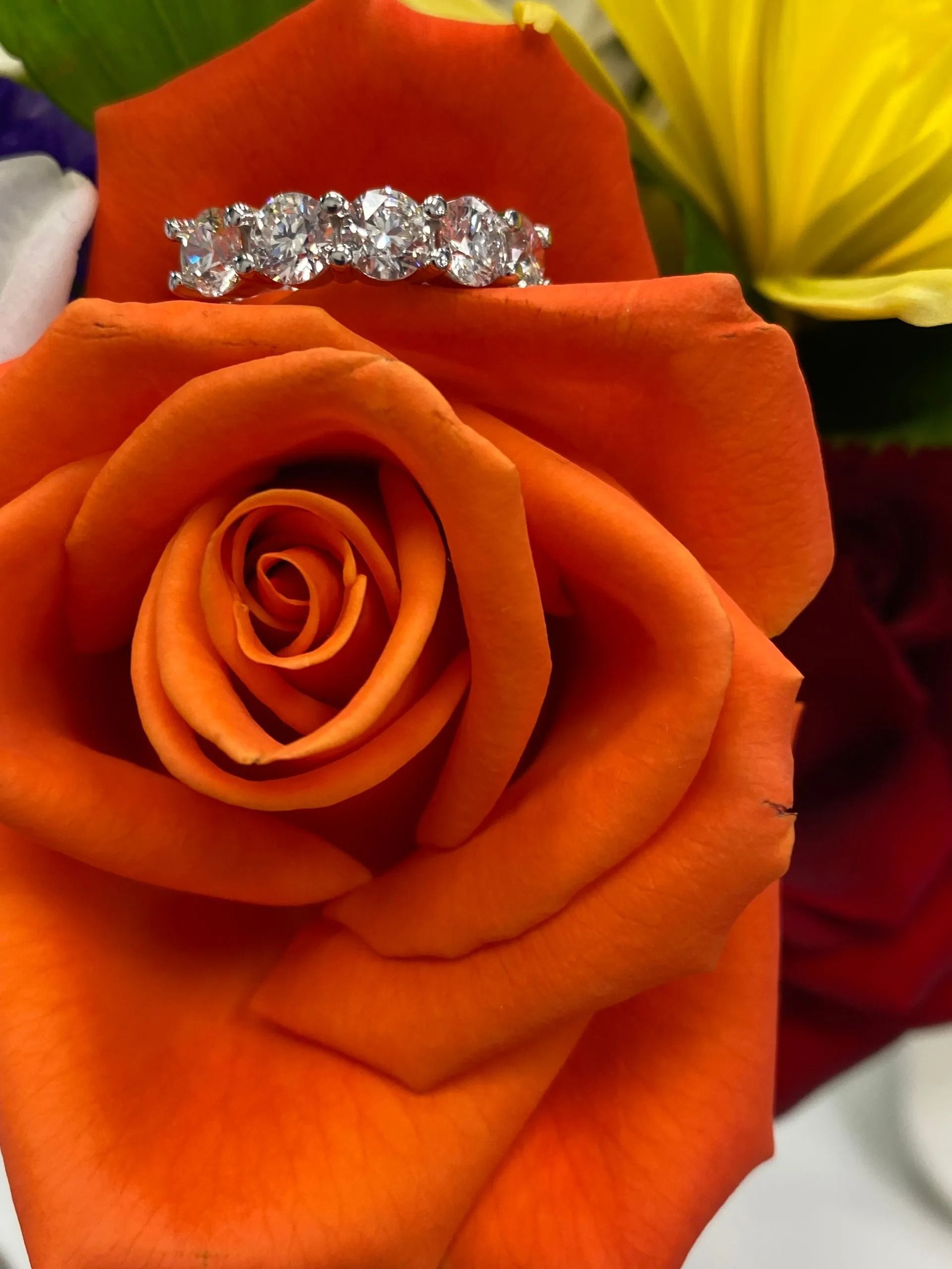 Orange rose with a diamond ring resting on its petals, against a colorful floral background