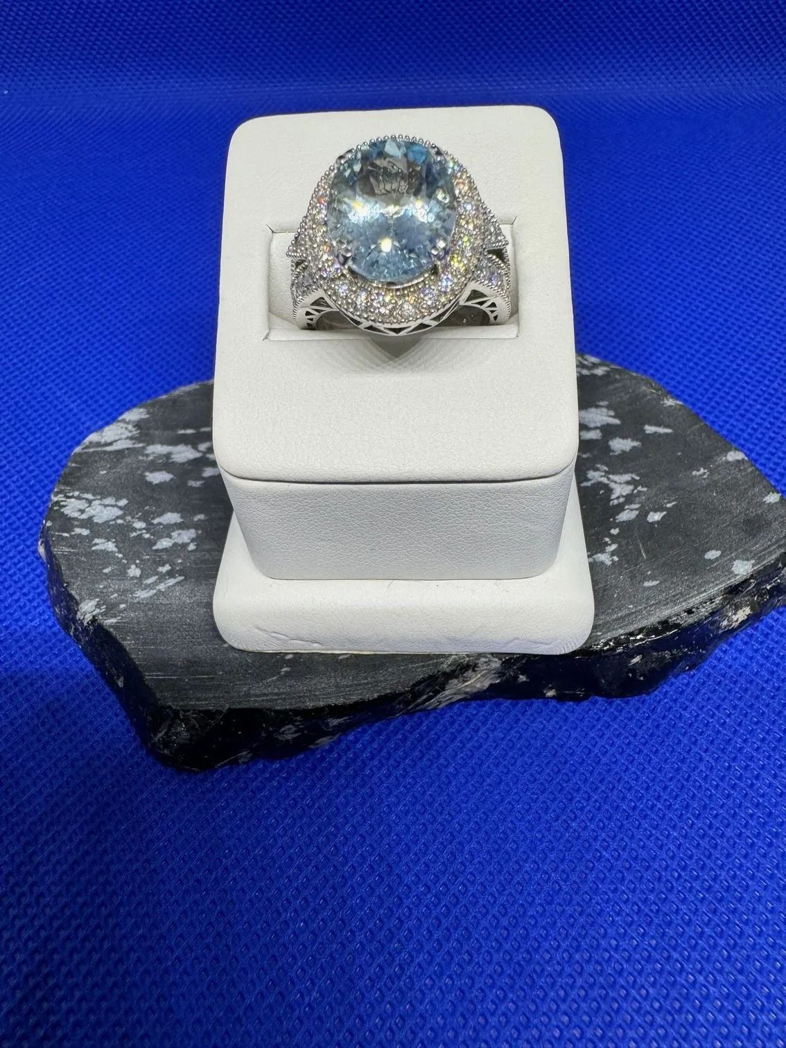Ring displayed on a white stand atop a dark stone base against a blue background