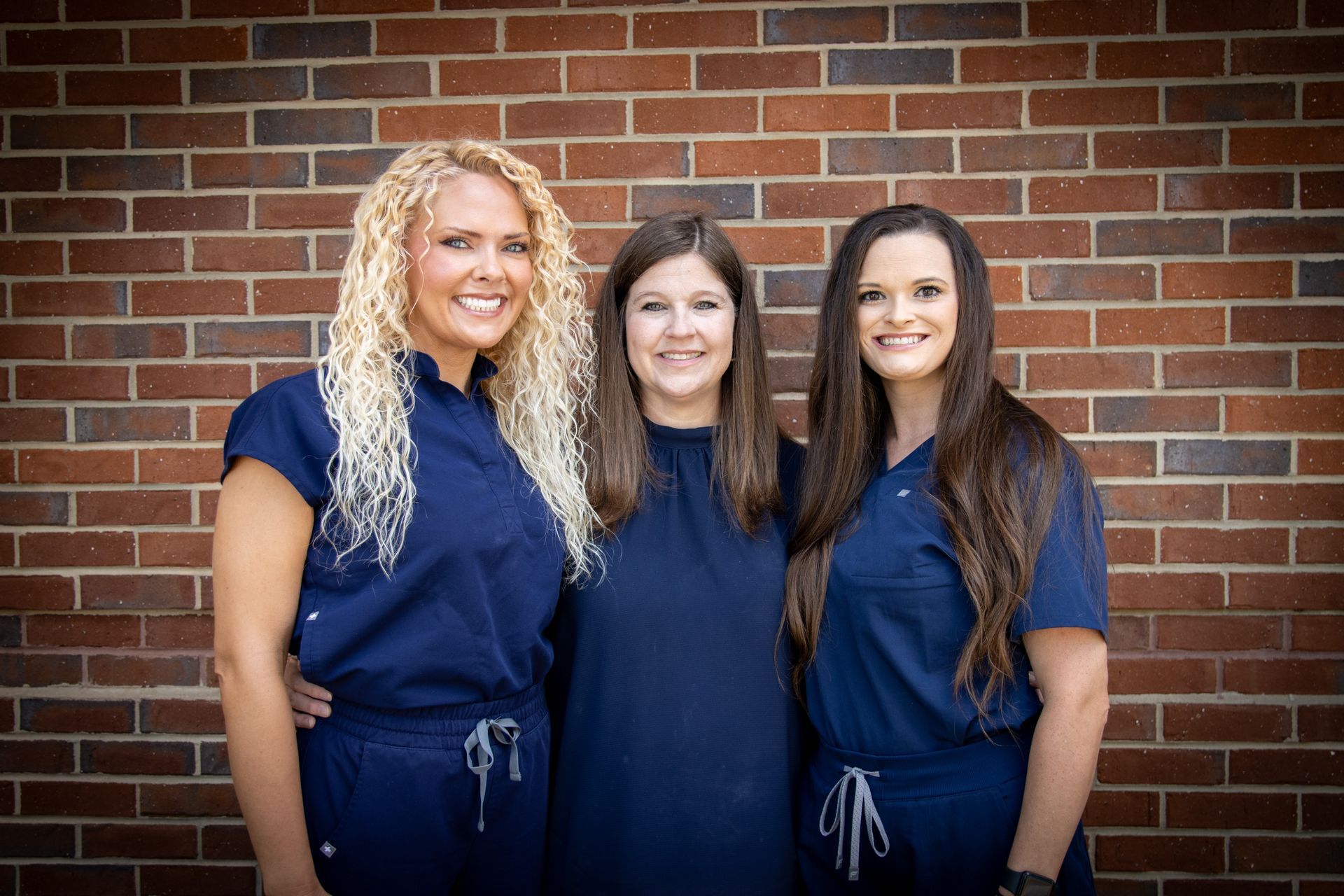 Three women in blue scrubs and dress posing in front of a brick wall.
