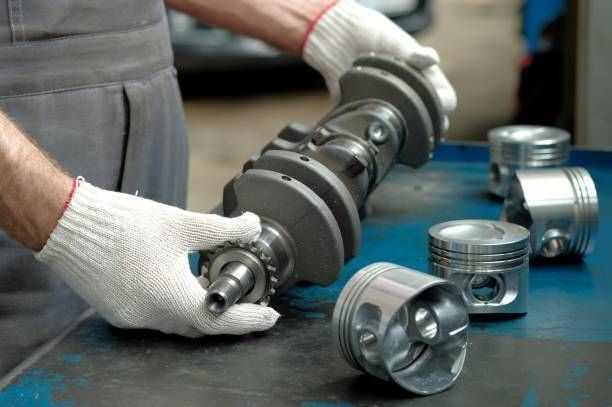 Hands wearing gloves holding a crankshaft with pistons in a workshop.