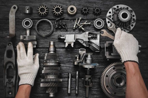 Hands in gloves, wrench, and various metal mechanical parts on a dark wooden surface.