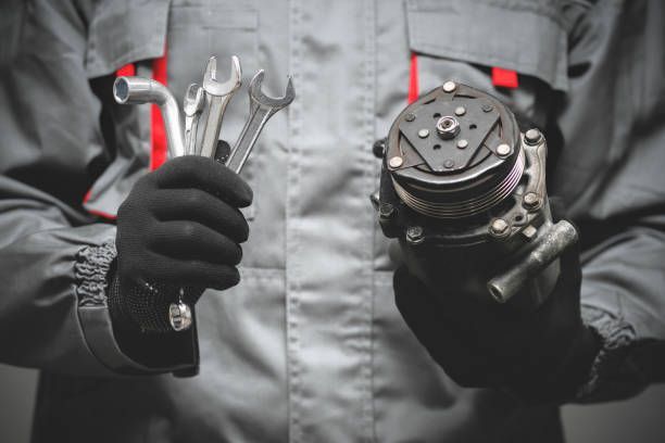 Mechanic in gray jumpsuit holding wrenches and a car part, both hands gloved.