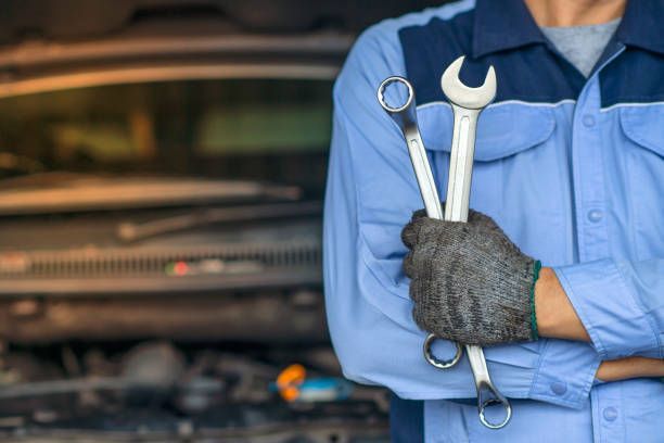 Mechanic holding wrenches, standing in front of an open car hood.