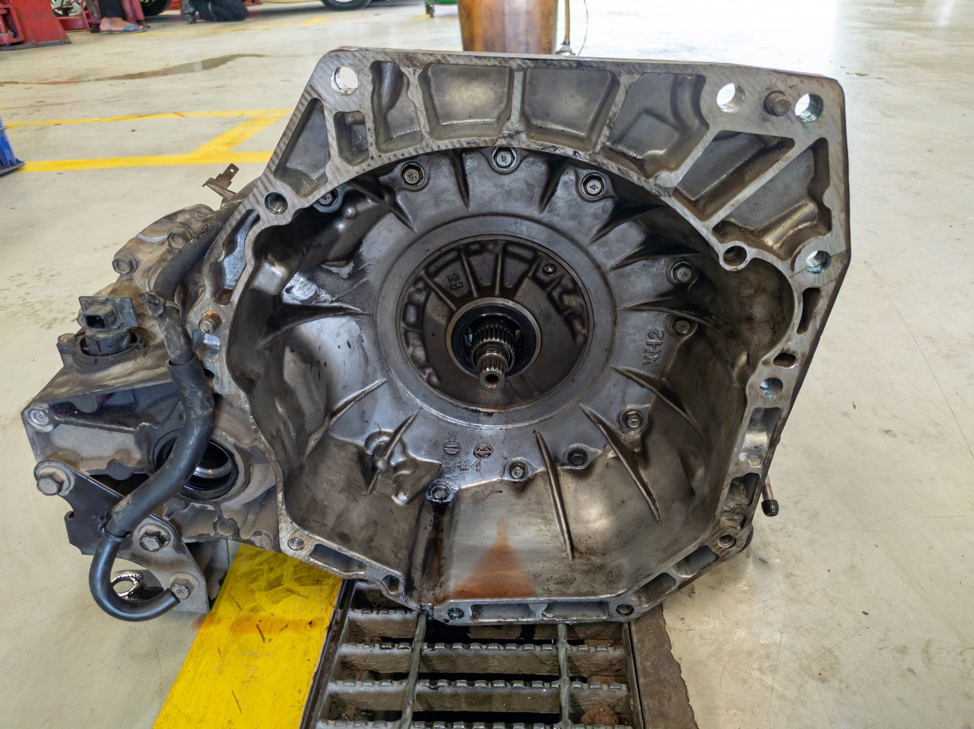 Open view of an automatic transmission housing on a shop floor. Gray metal with visible oil stains, dark inner components.
