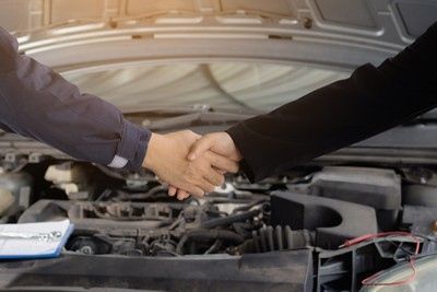 Hands shaking in front of an open car hood, possibly a mechanic and customer, sealing a deal.