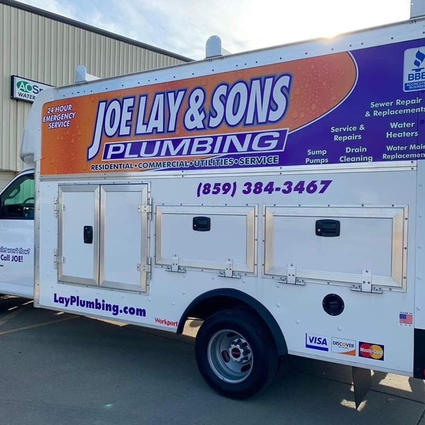 Joe Lay & Sons Plumbing truck parked outside a building. White with company logo, phone number, and services listed.