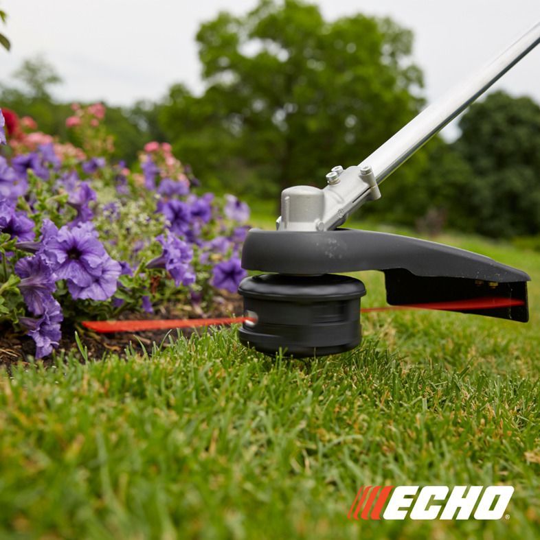 A lawn mower is cutting a lush green lawn with purple flowers in the background.