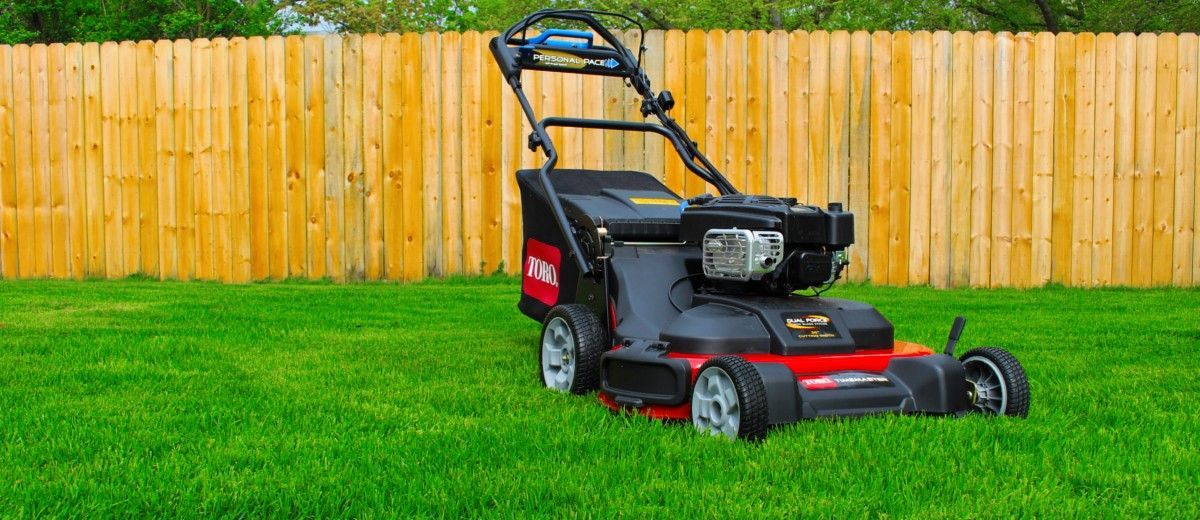 A lawn mower is sitting on top of a lush green lawn in front of a wooden fence.