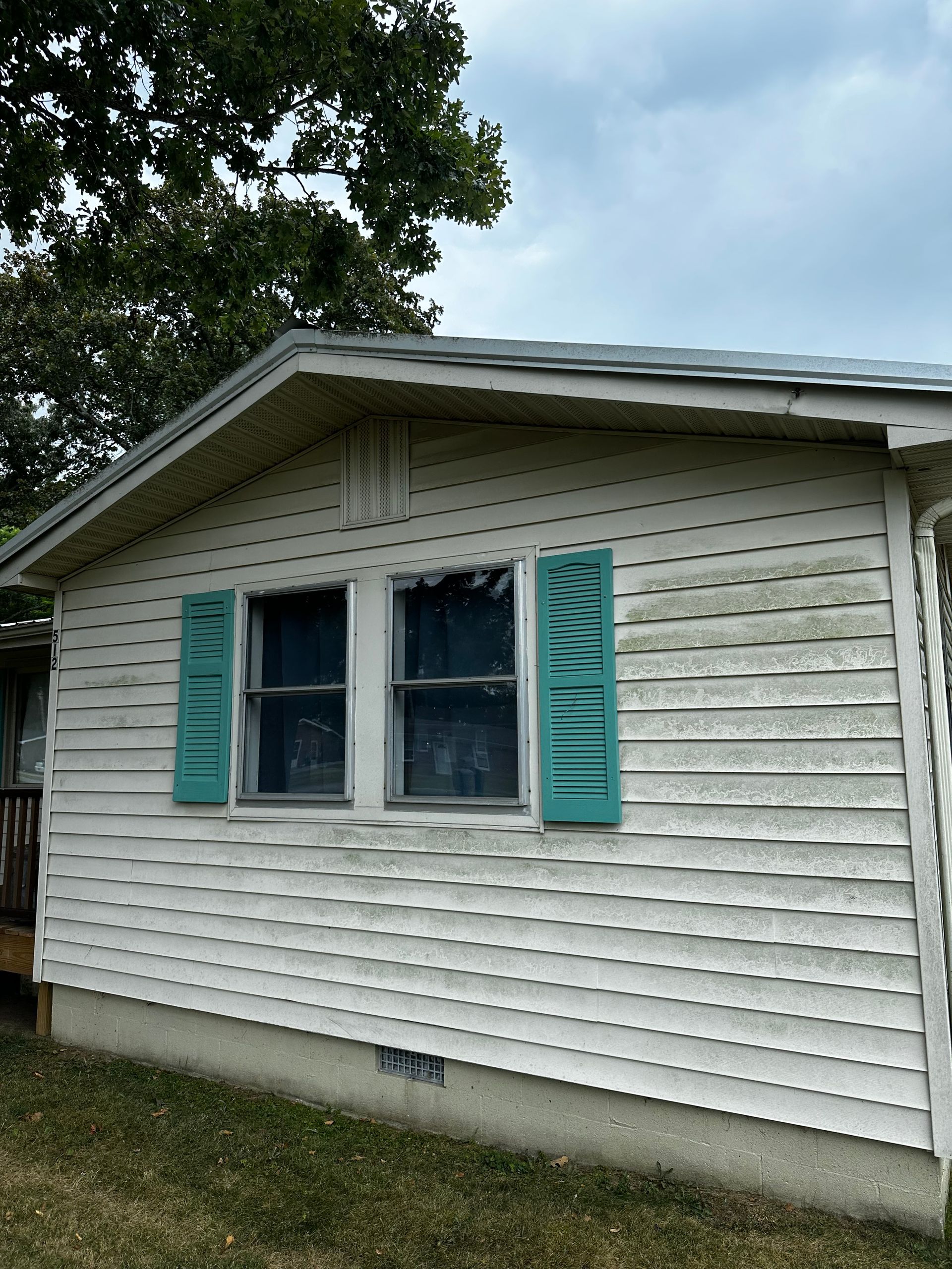 White house with turquoise shutters and moldy siding under cloudy sky.