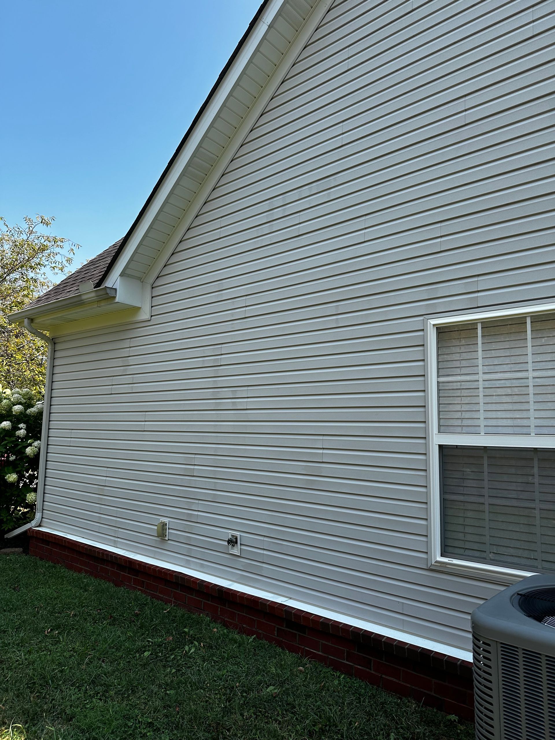 Exterior view of a light gray house with white trim, red brick foundation, and green grass. A window and AC unit are visible.