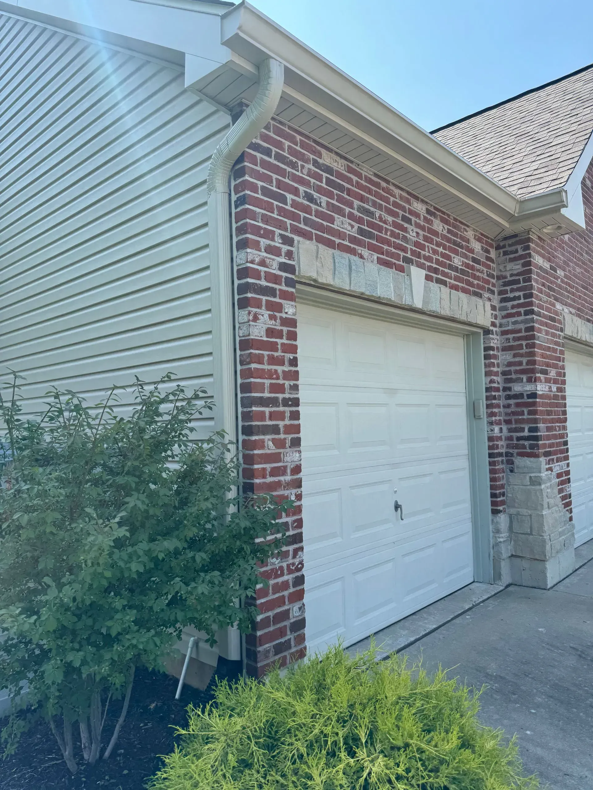 Brick house exterior with a white garage door, beige siding, and shrubs in front
