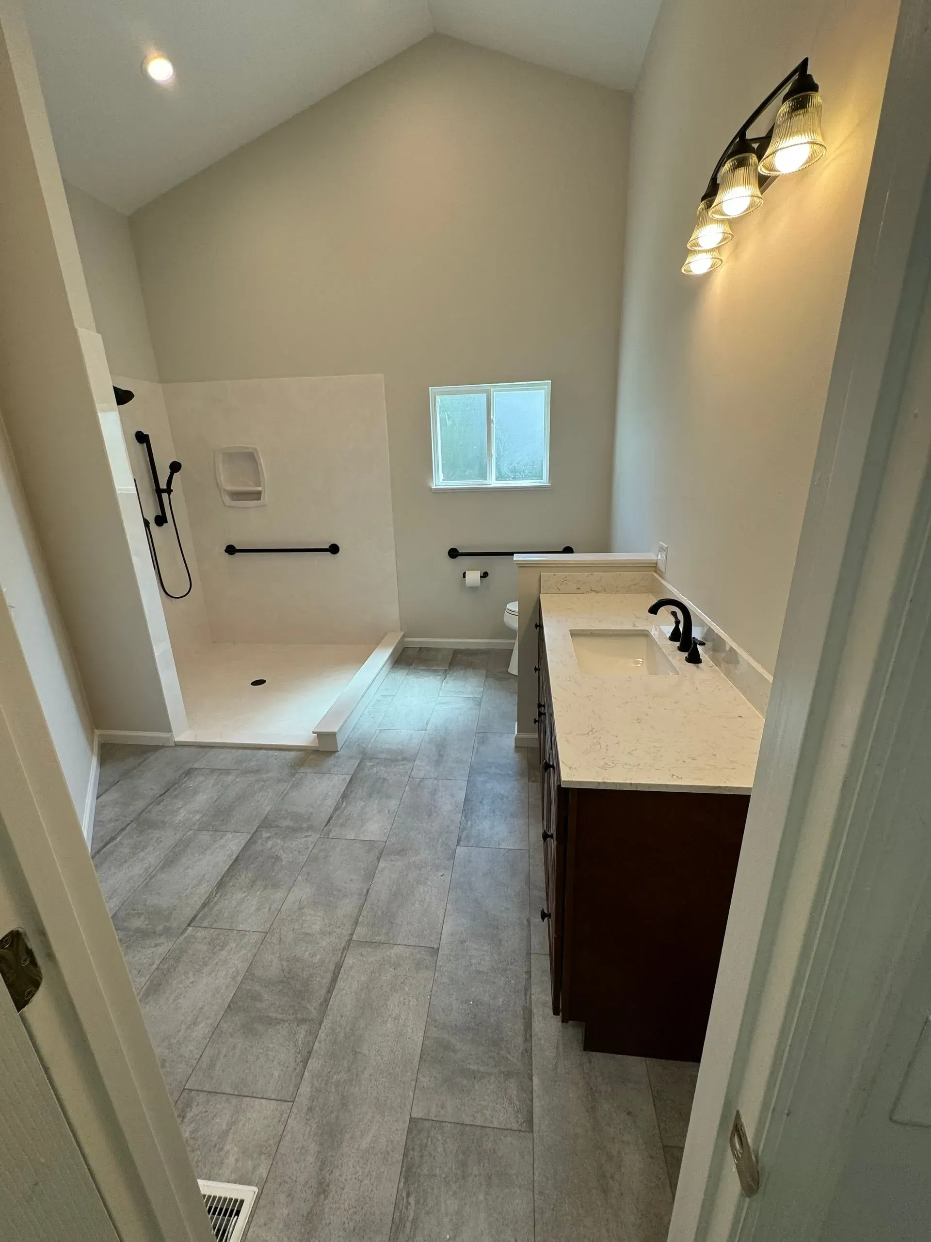 Bathroom with shower, vanity, wall sconce, and gray tile floor in a bright neutral room