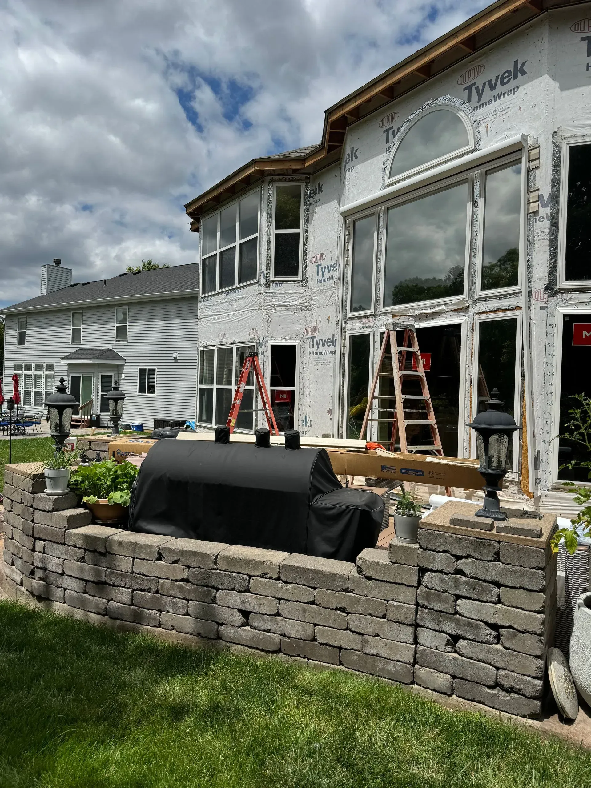 House exterior under renovation with ladders, windows, and a stone retaining wall in front of a grassy yard