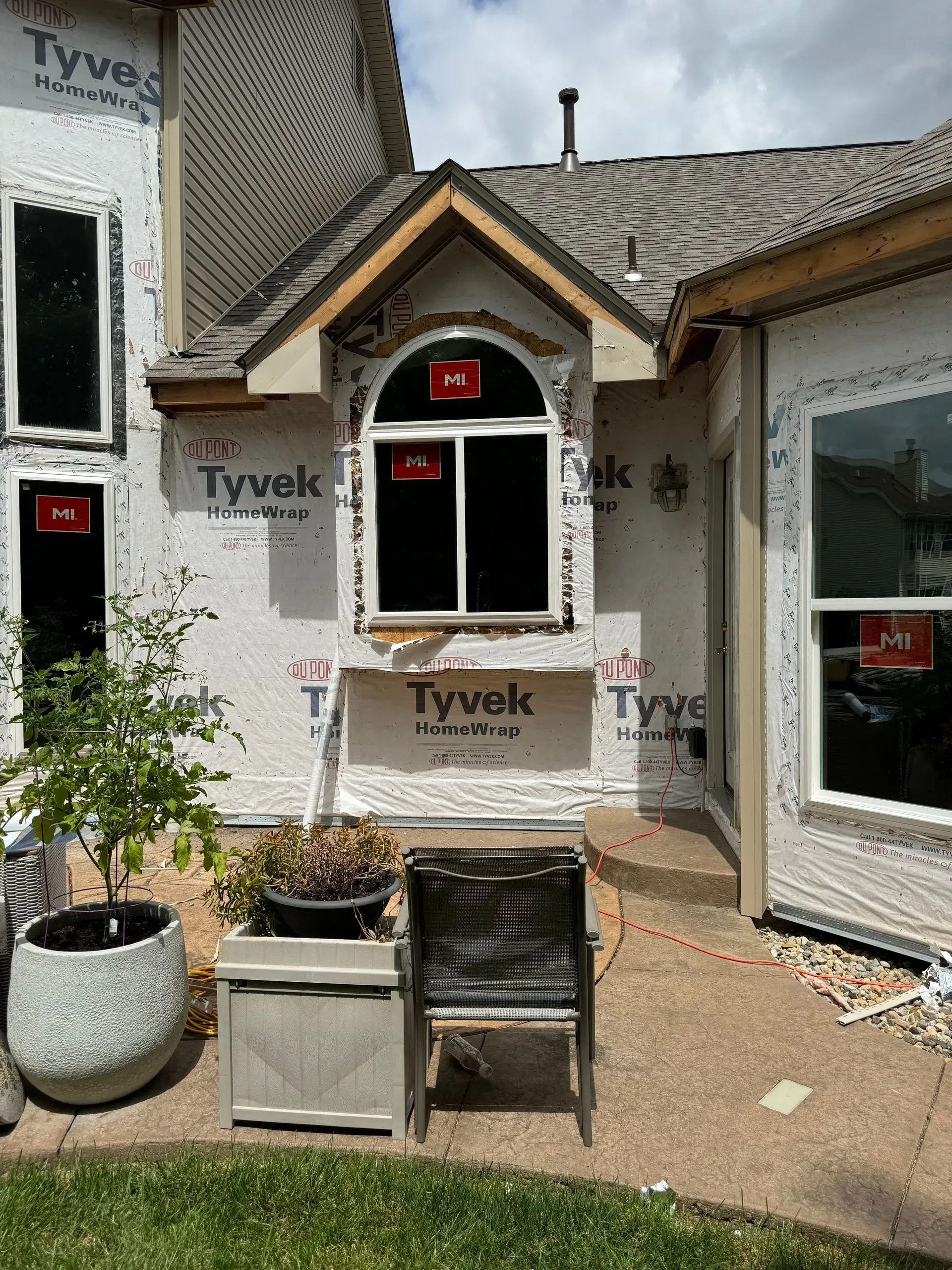 Small house exterior under renovation with Tyvek wrap, arched window, and patio furniture in front