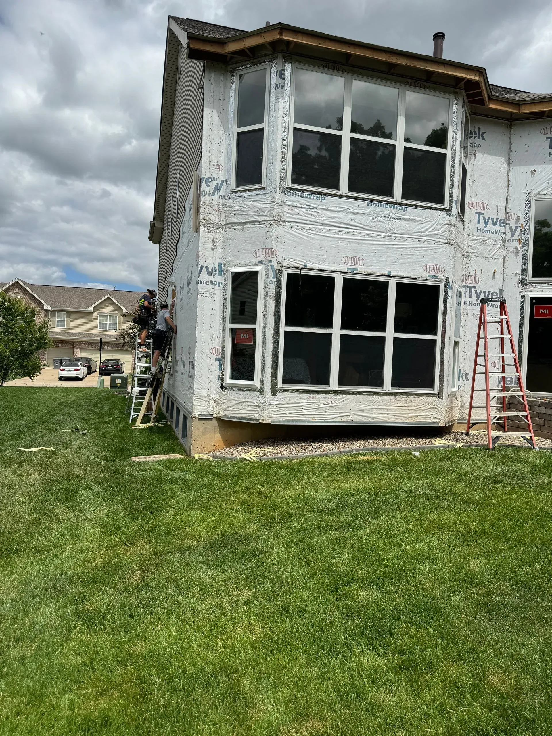 Partially renovated house exterior with peeling white paint, large windows, and a red ladder on the lawn