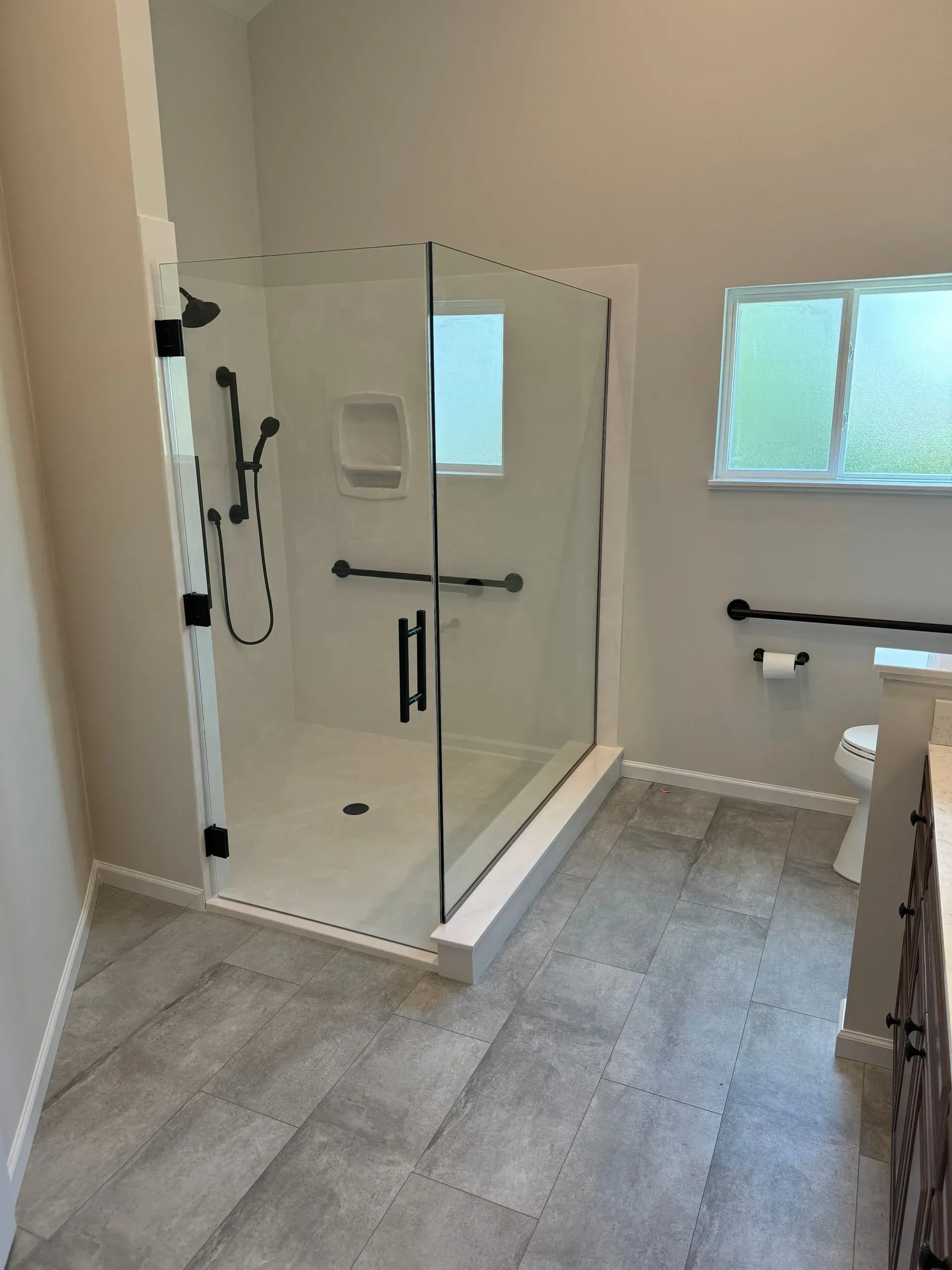 Modern bathroom with glass walk-in shower, gray tile floor, and frosted window.