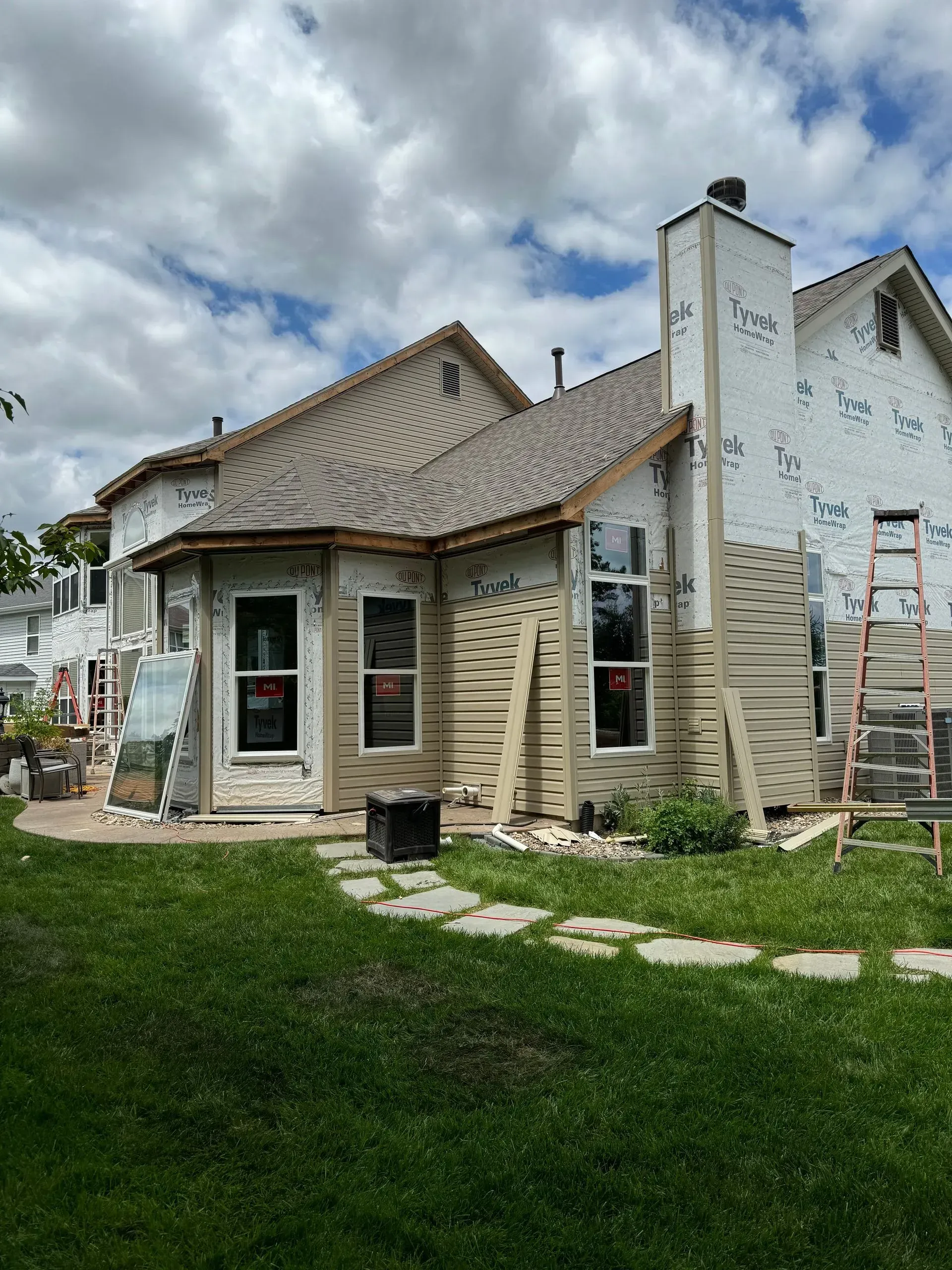 Single-story house under renovation with ladders, stone facade, and a green lawn in front.
