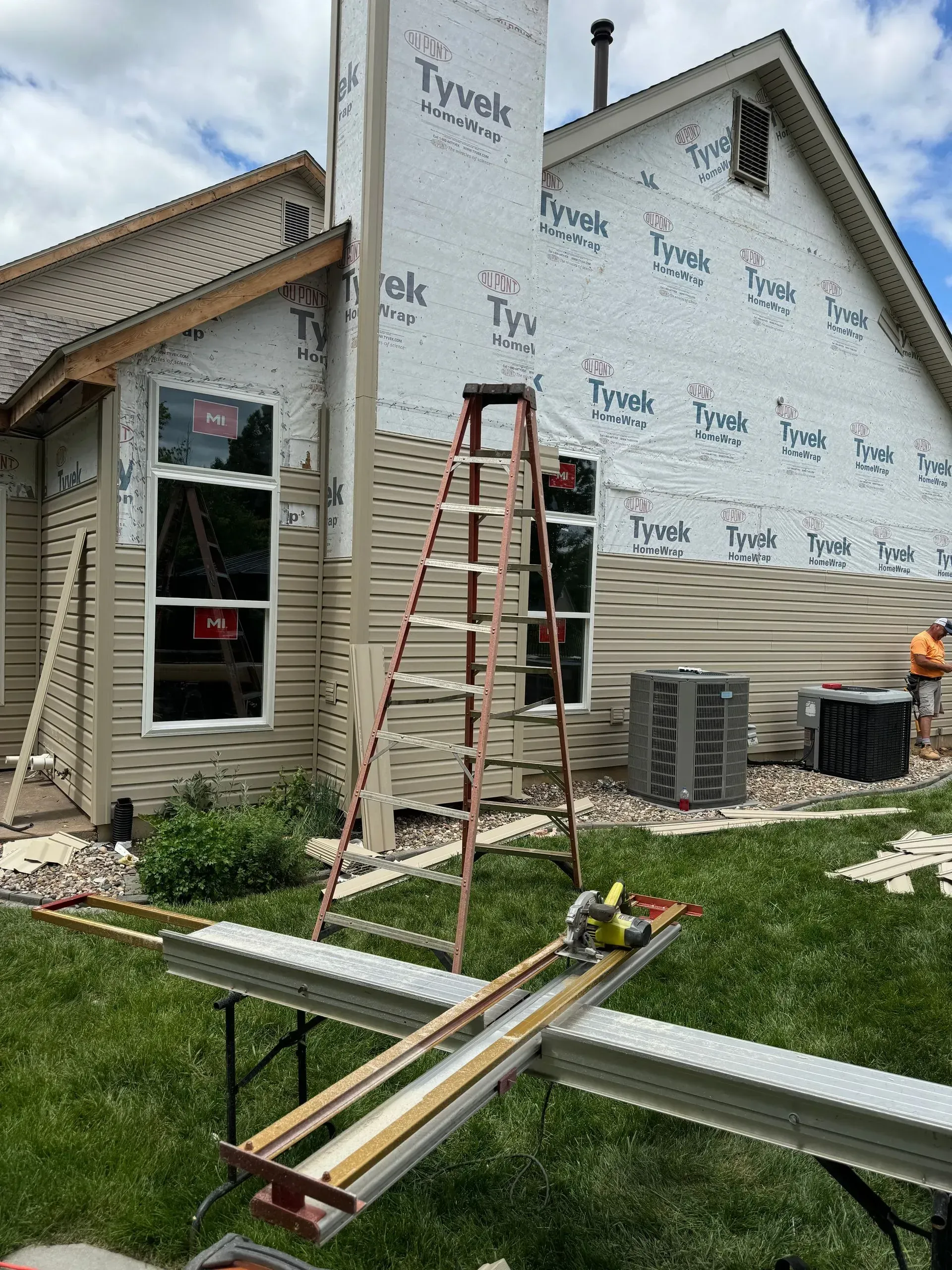 House exterior under construction with ladders, scaffolding, and siding wrapped in Tyvek.
