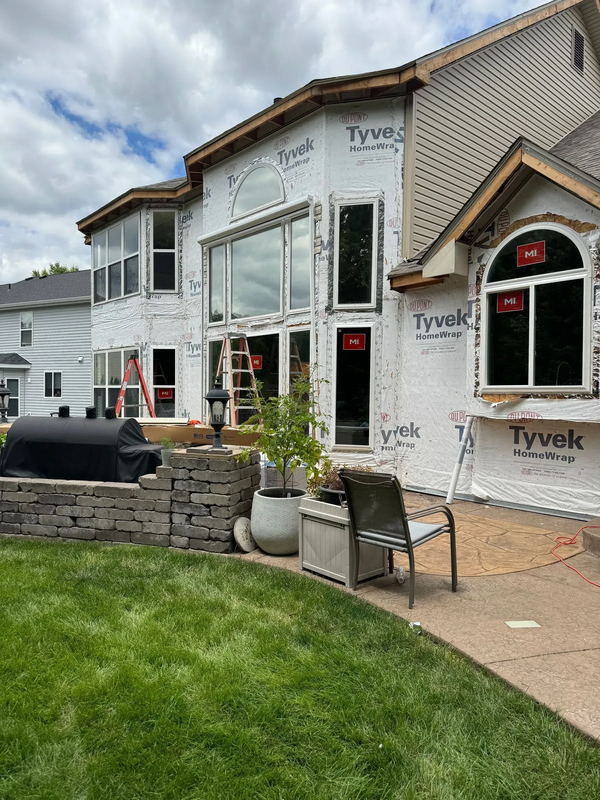 Partially built house with Tyvek wrap, large windows, stone wall, and a green lawn under a cloudy sky
