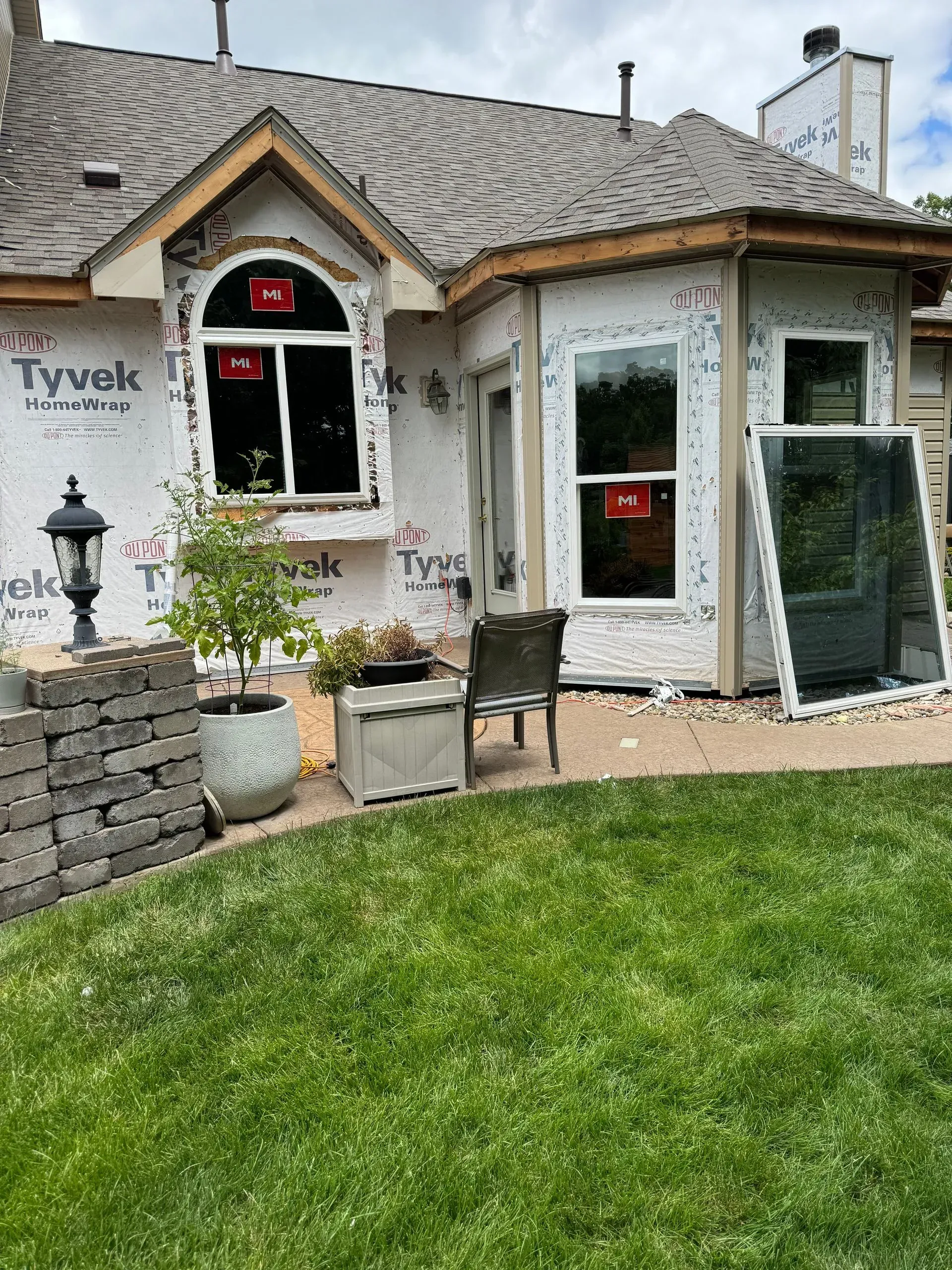 Partially renovated house exterior with Tyvek wrap, white-trimmed windows, stone wall, and green lawn
