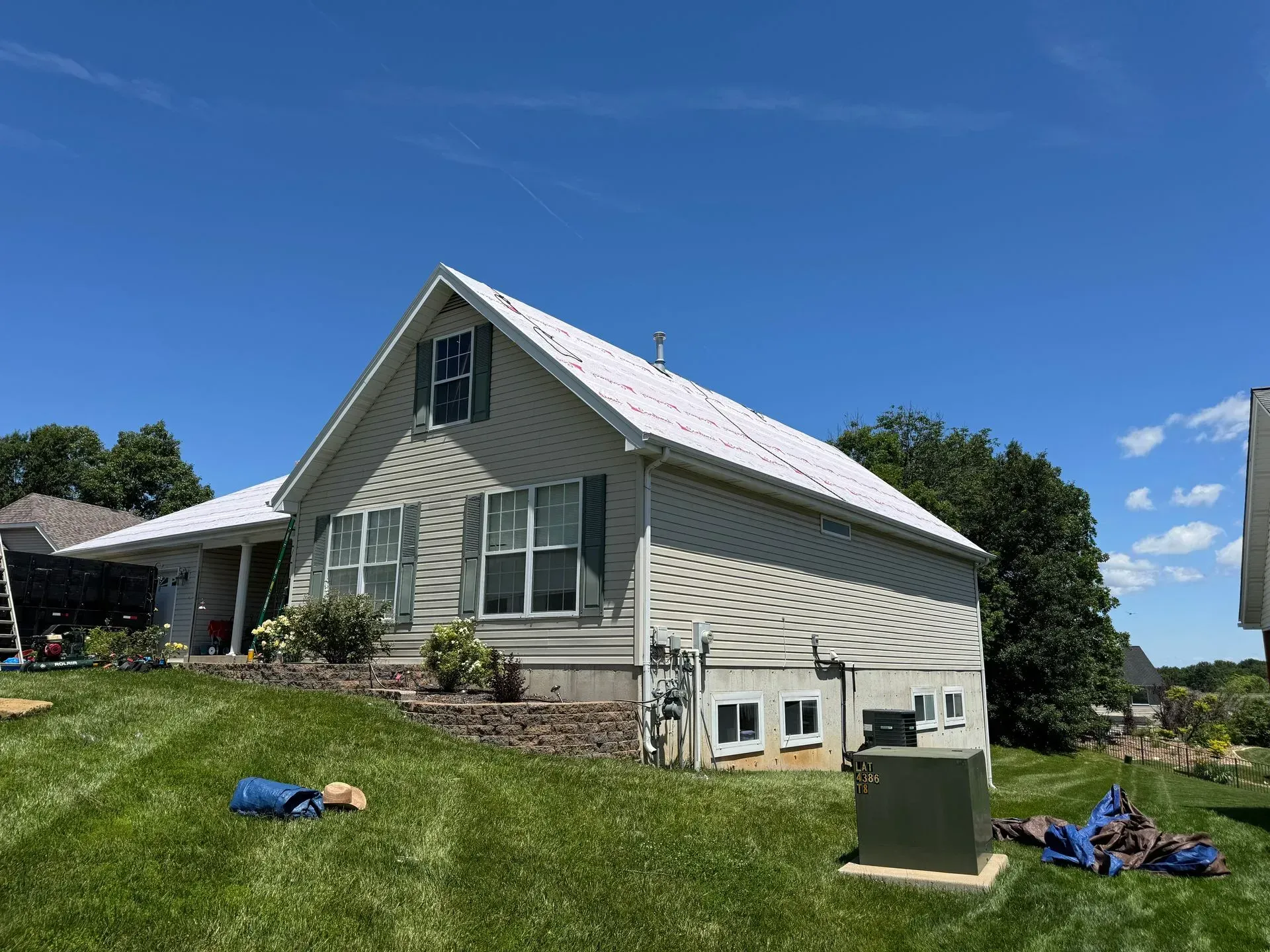 White house with gray roof on a grassy hill under a blue sky, with yard equipment in front.