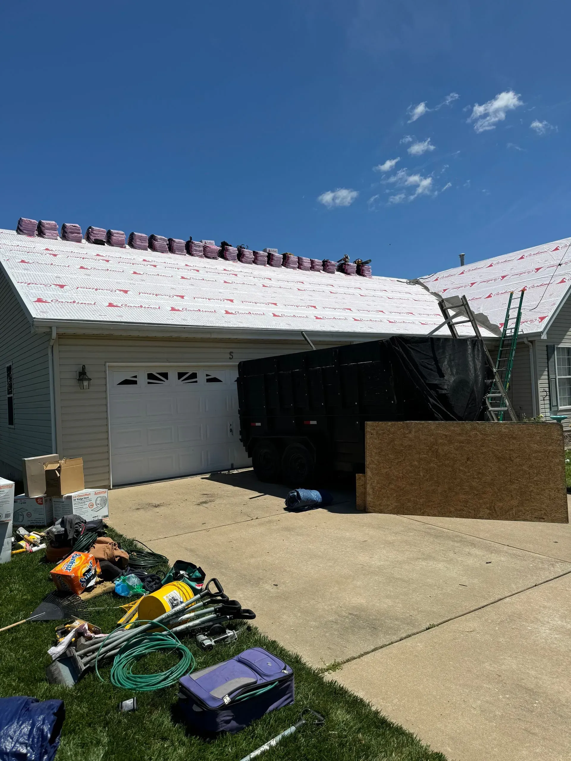 Damaged house roof with exposed underlayment, debris scattered in the yard, and a garage in view.