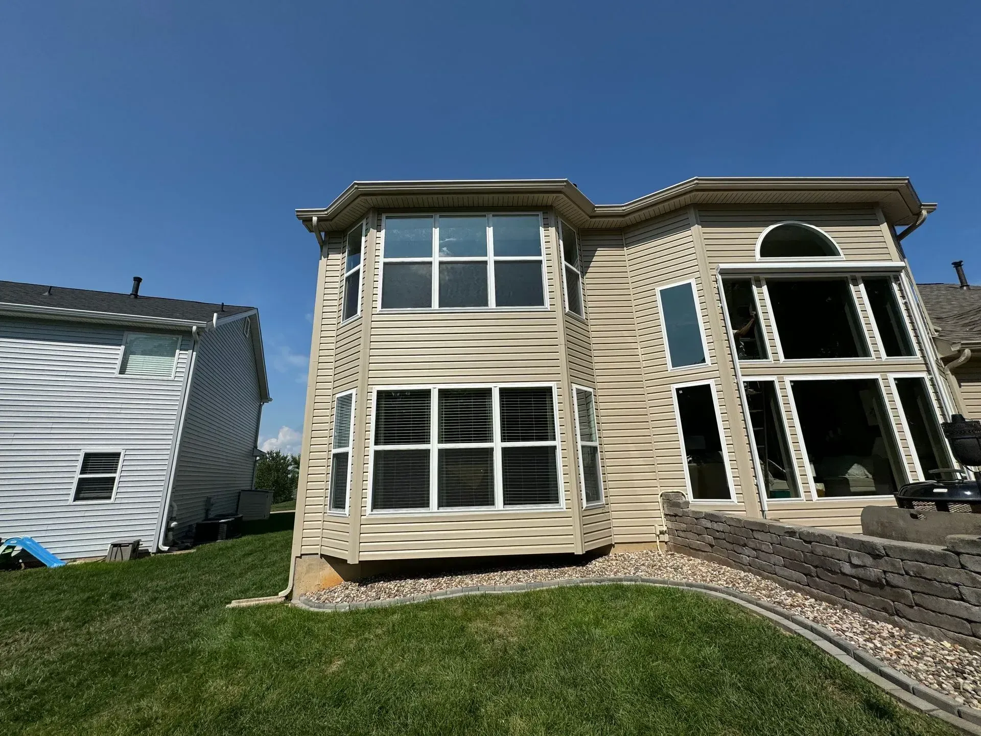 Two-story beige house with large windows, stone accents, and a grassy front yard under a blue sky
