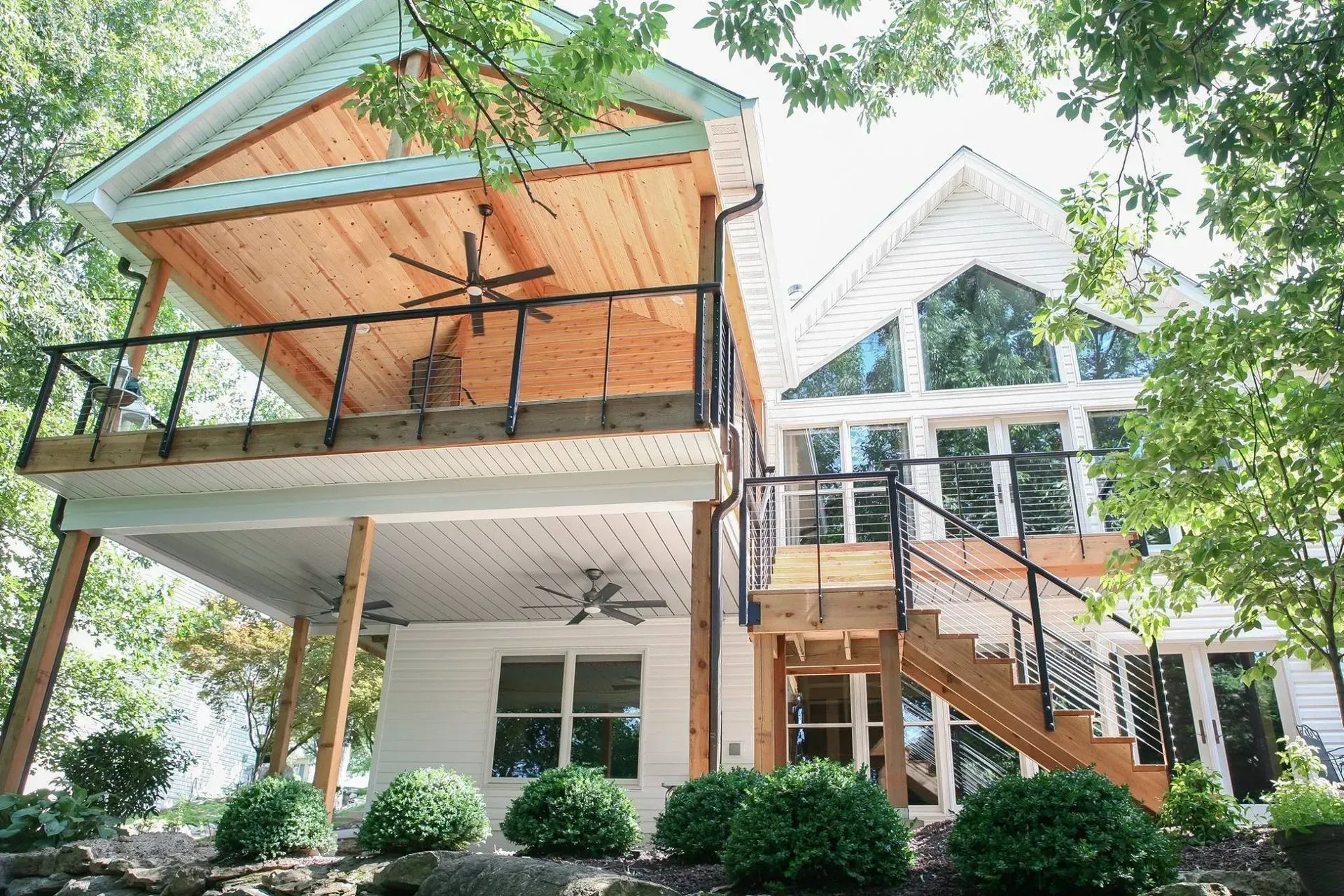 Modern house exterior with large windows, balconies, and stairs surrounded by trees and shrubs