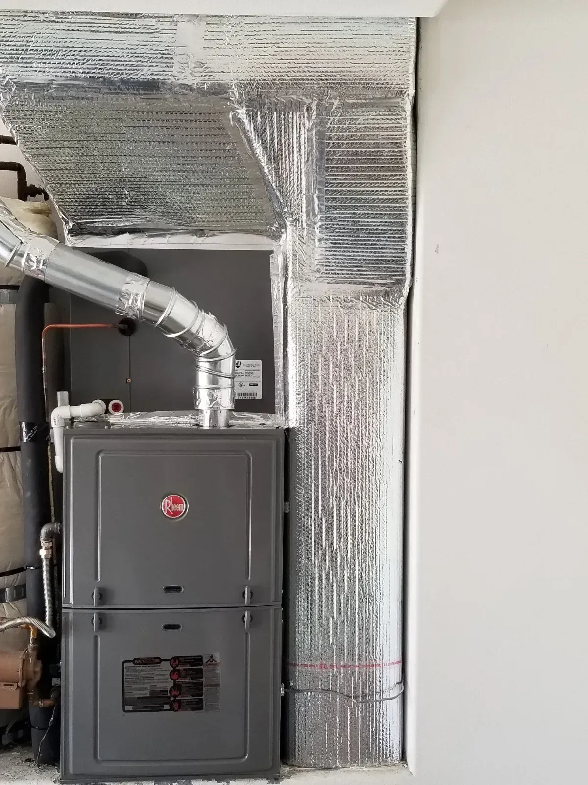 Furnace unit with ductwork, wrapped in silver insulation, in a utility closet.