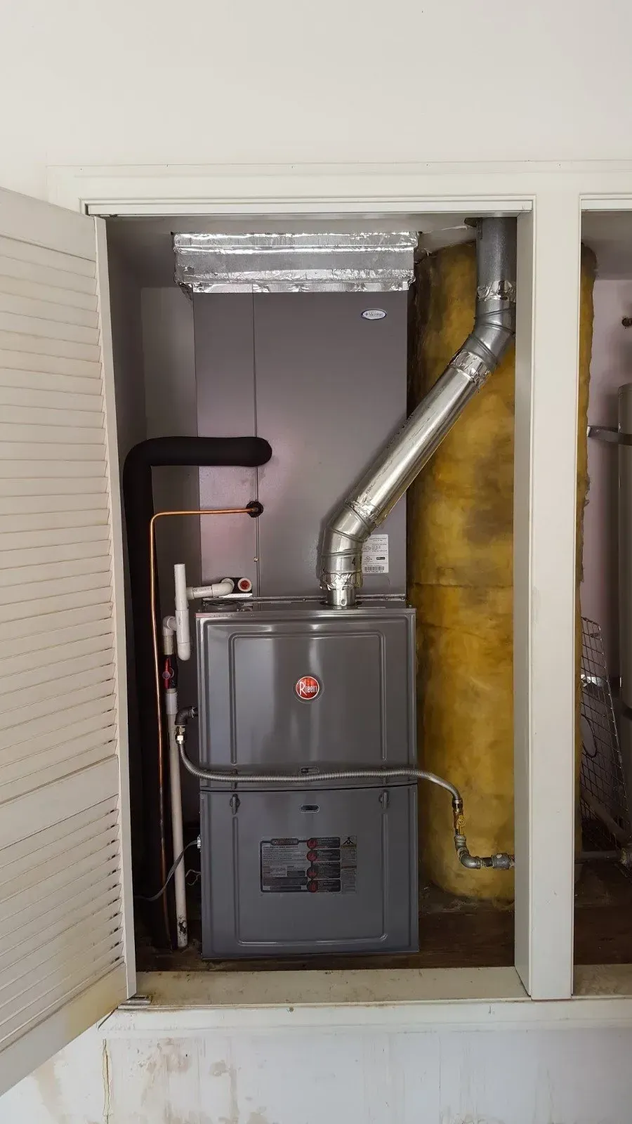 A furnace unit inside a closet with ductwork and a vent pipe. The unit is gray and silver.
