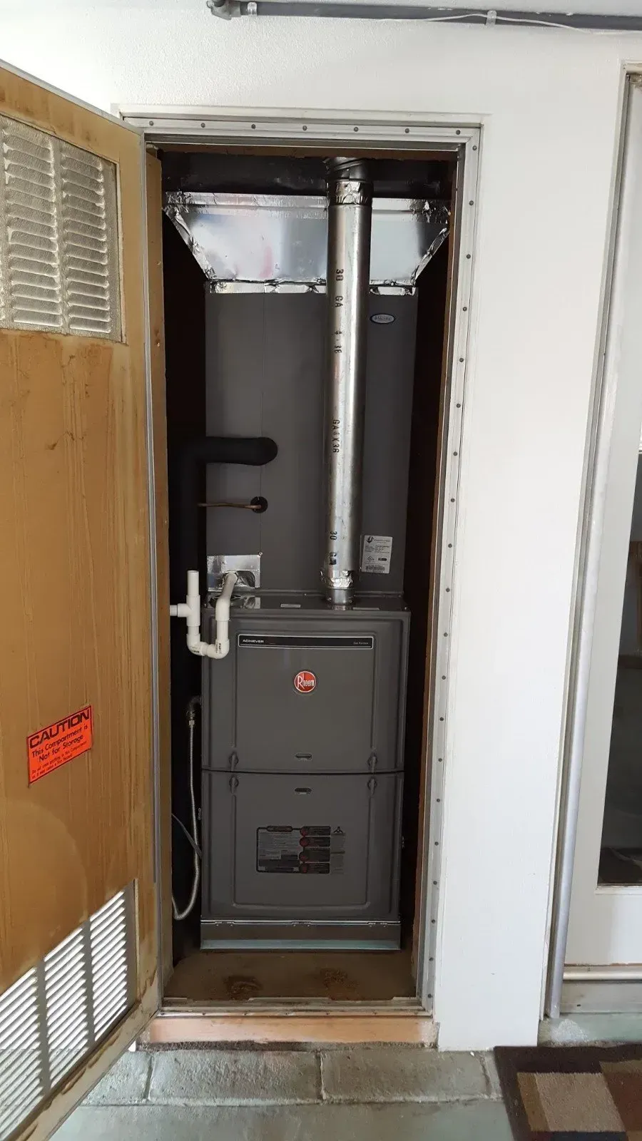 A vertical furnace, in a doorway, with silver exhaust pipe. A metal door with vents is on the left.